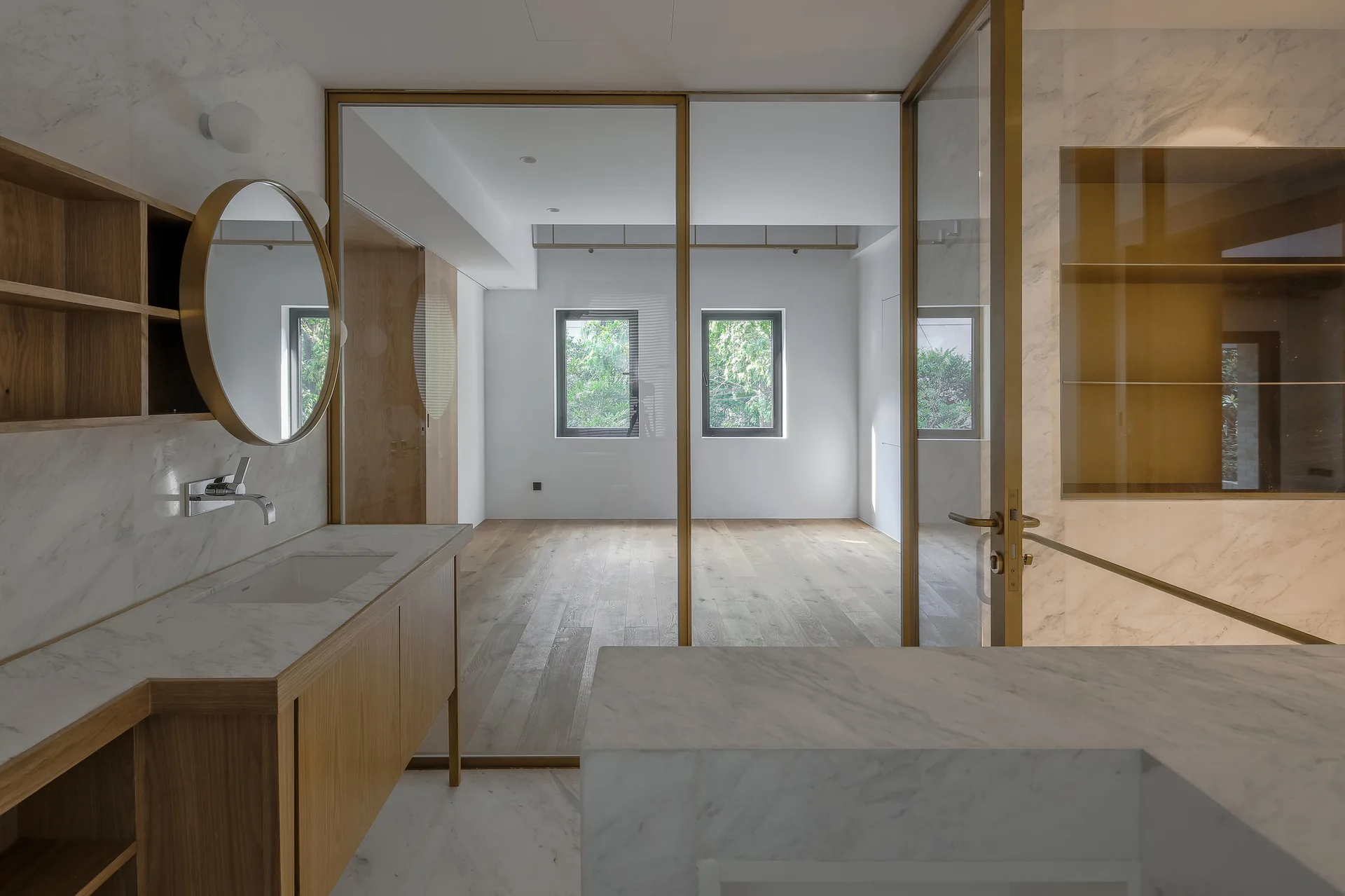 A minimalist bathroom featuring a marble vanity, round brass mirror, and a glass partition wall looking into a wood-floored room.