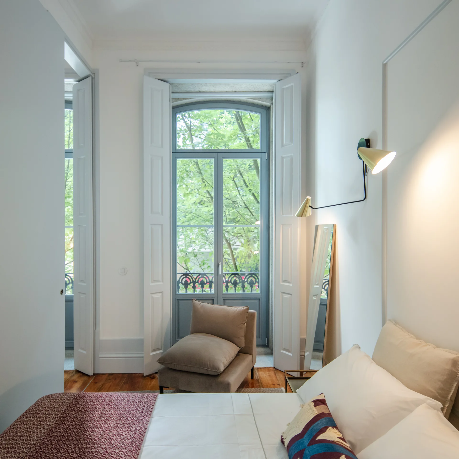A bright bedroom featuring a white bed, a brass wall sconce, and a tan lounge chair beside a tall shuttered window.