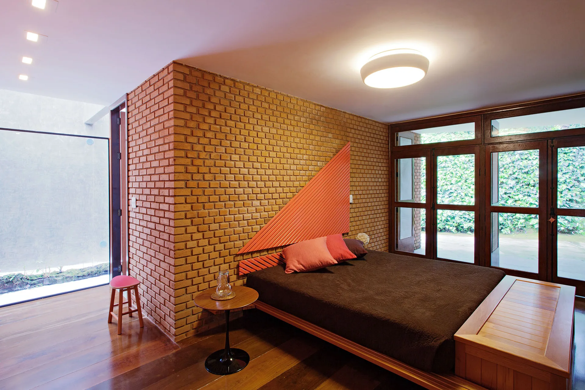 A contemporary bedroom features an exposed red brick wall, a platform bed with a slatted triangular coral headboard, and wood-framed glass doors.