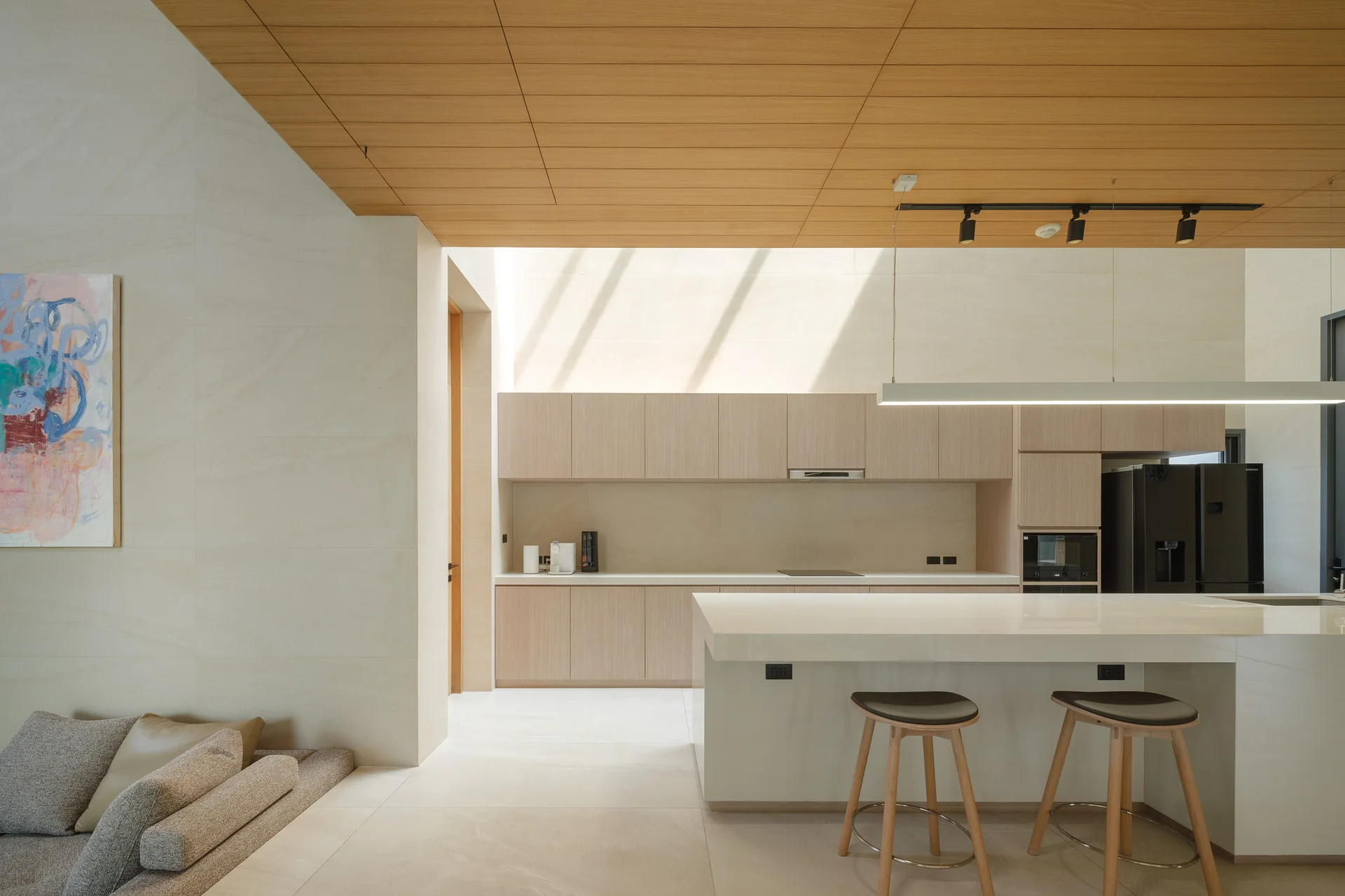 A minimalist kitchen features light wood cabinets, a large white island with two stools, and a warm wood plank ceiling.