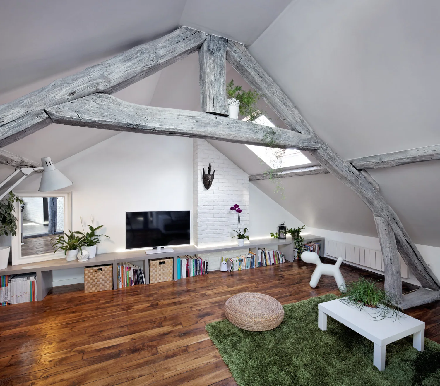 An attic living room features weathered timber beams, a low grey media console beneath a sloped ceiling, and a green shag rug.