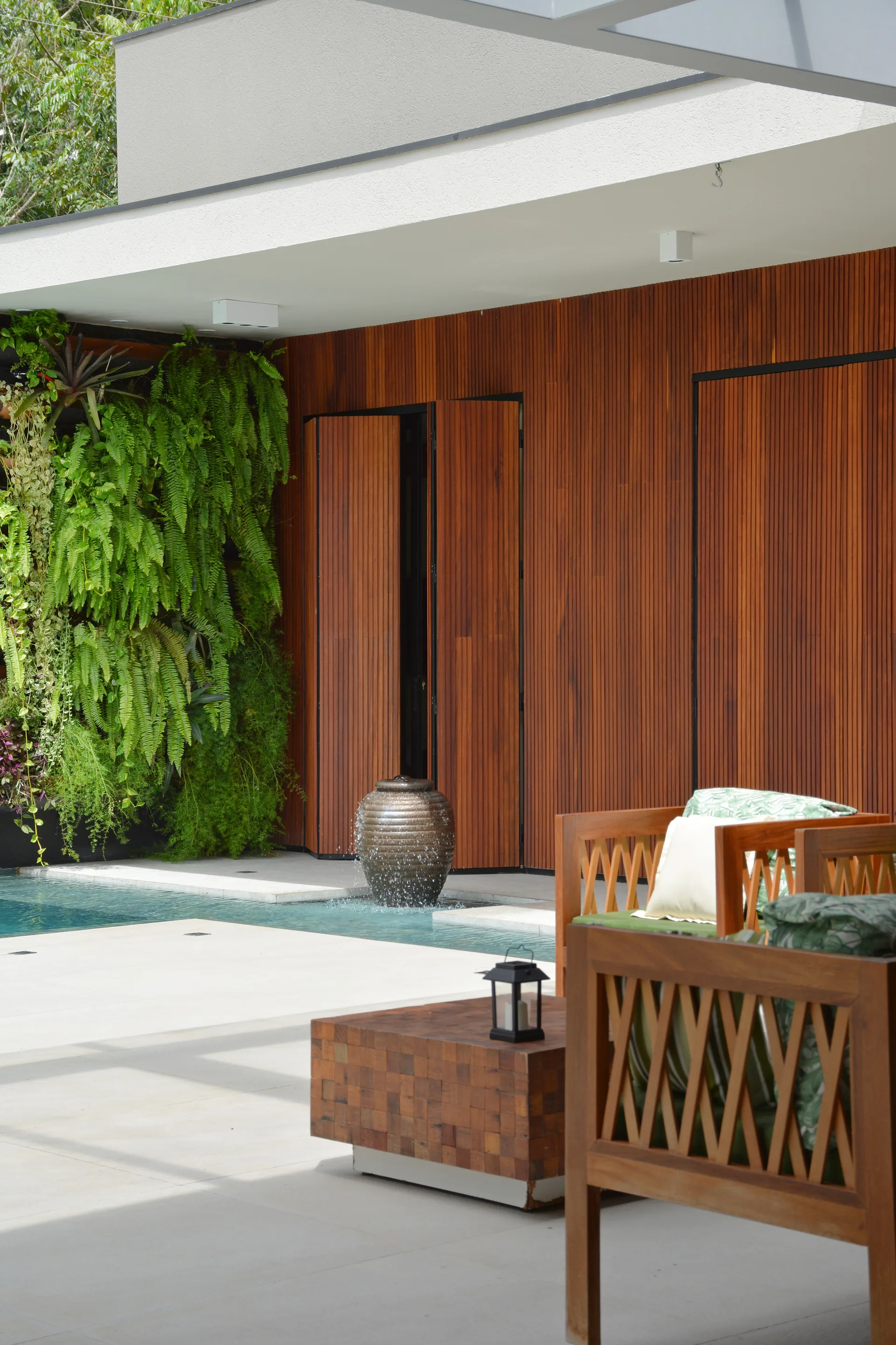 An outdoor patio features wooden lattice armchairs, a block-style coffee table, a water fountain, and a lush vertical garden wall next to a pool.