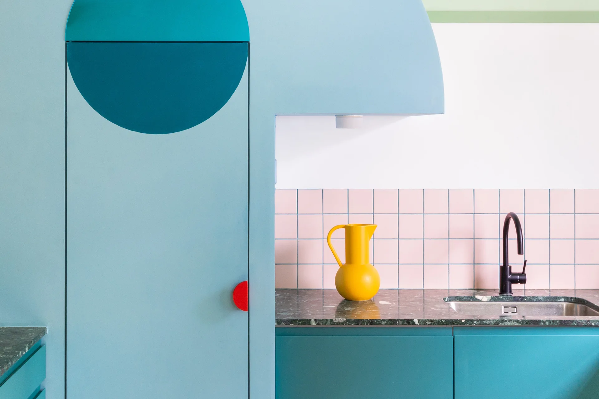 Blue kitchen cabinets with geometric cutouts stand beside a green stone countertop, pink square tile backsplash, and a black gooseneck faucet.