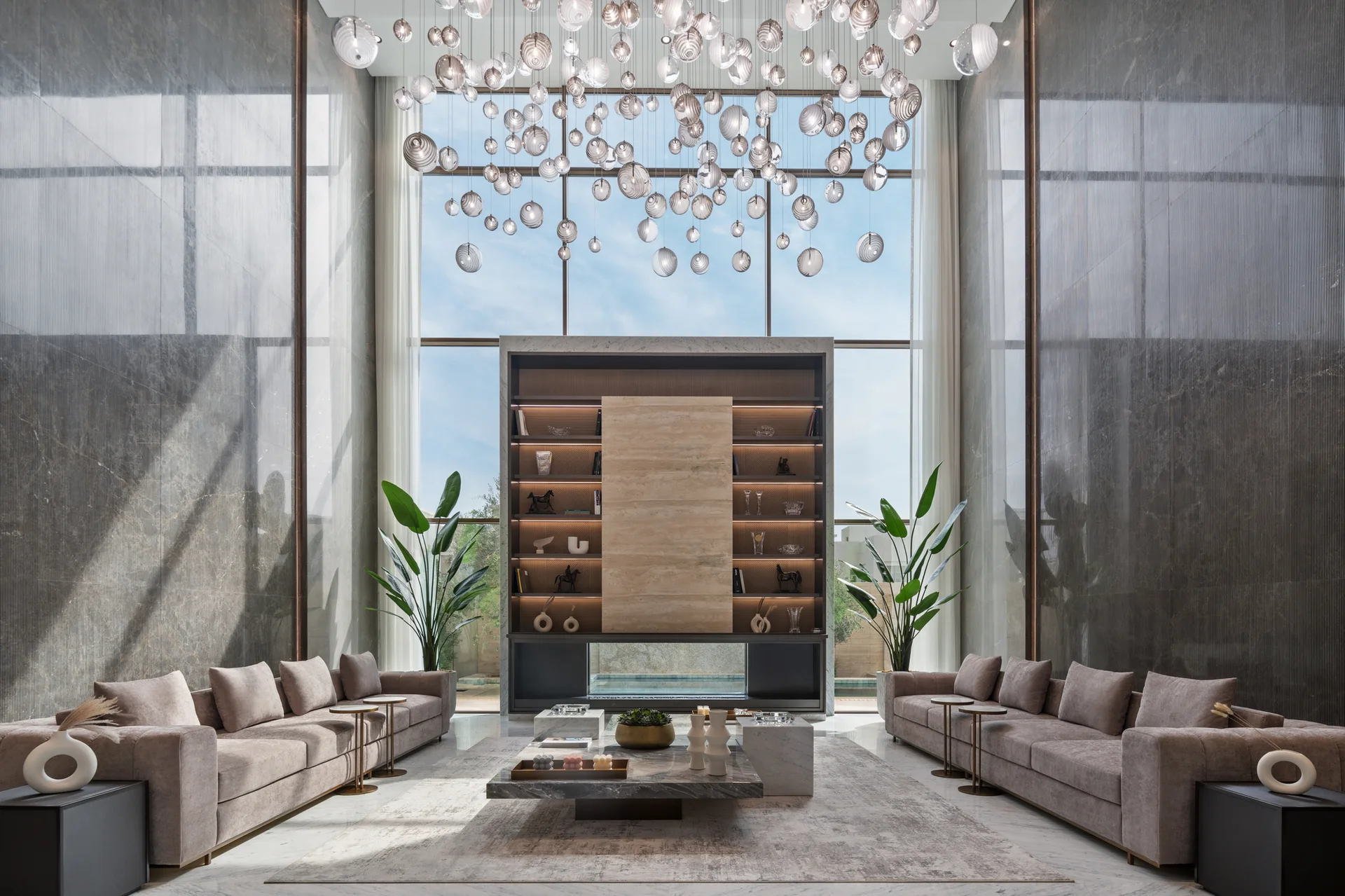 Two symmetrical sofas face each other in a double-height living room featuring grey marble walls and a massive cluster of glass pendant lights.