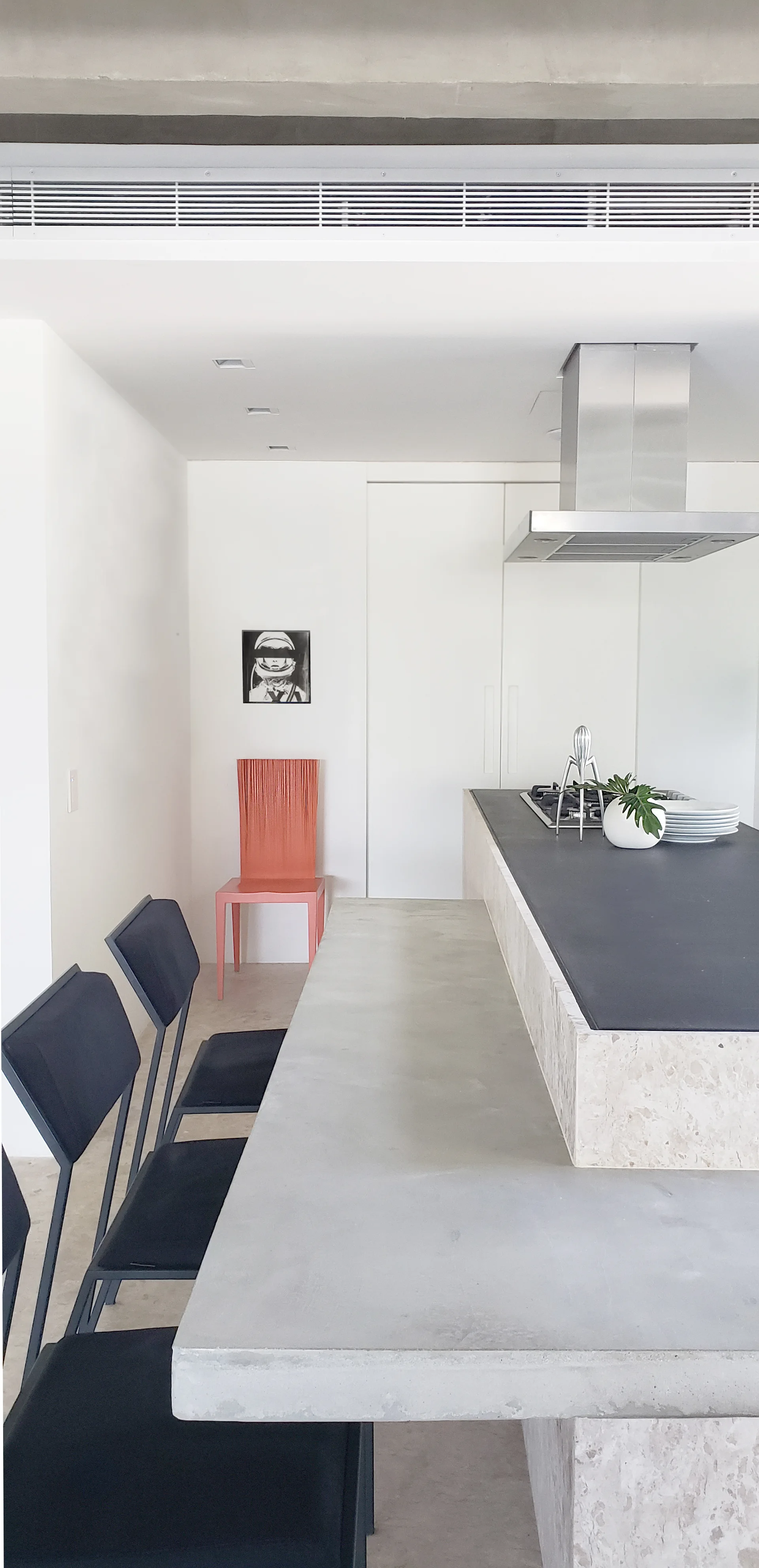 A minimalist kitchen features a split-level concrete and black island with matching chairs and a single coral chair in the background.