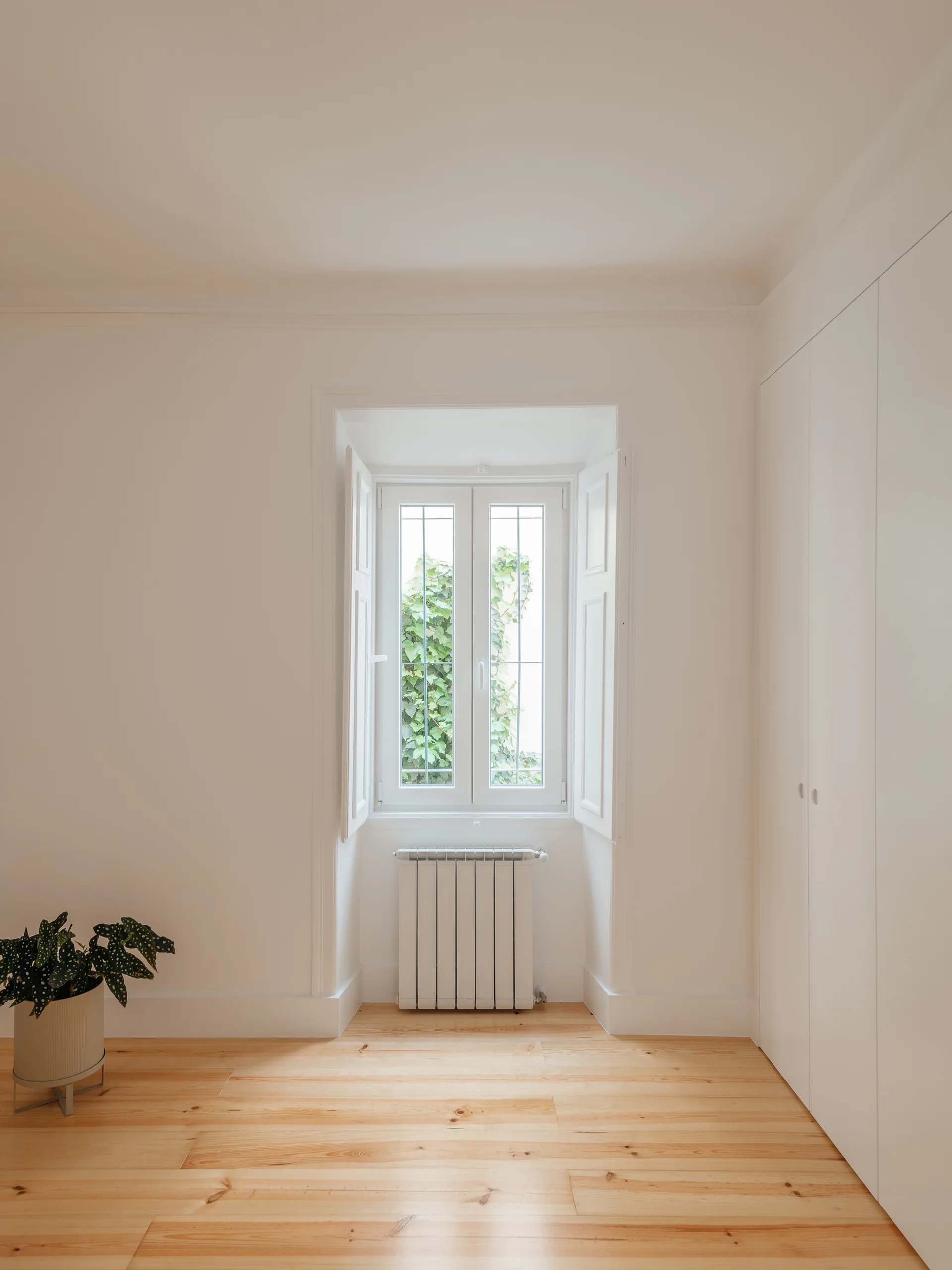 A minimalist white room featuring natural wood flooring, a recessed window with white shutters, and built-in closet doors.