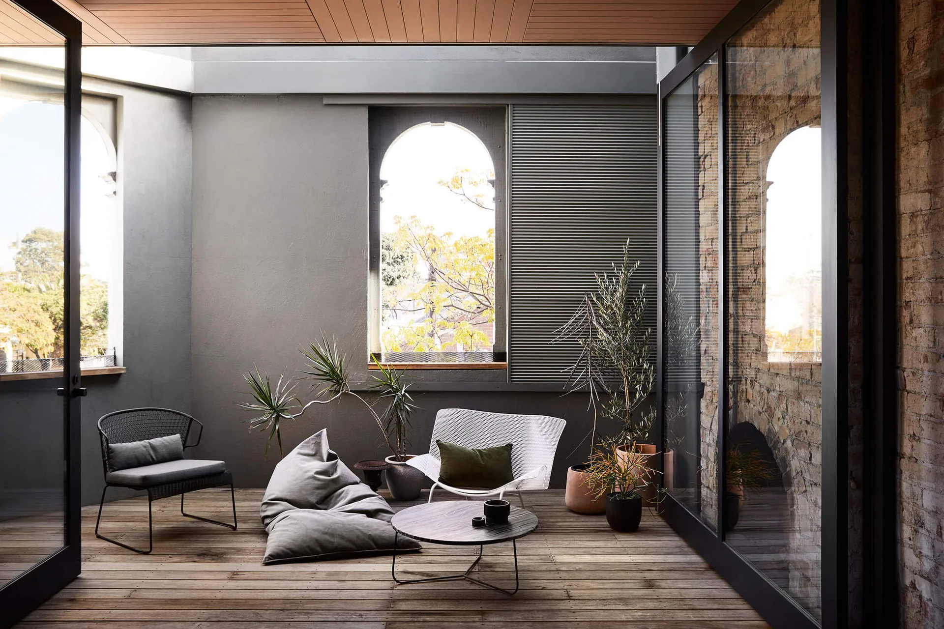 A covered patio with wood decking features modern lounge chairs, potted plants, an arched window, and an exposed brick wall.