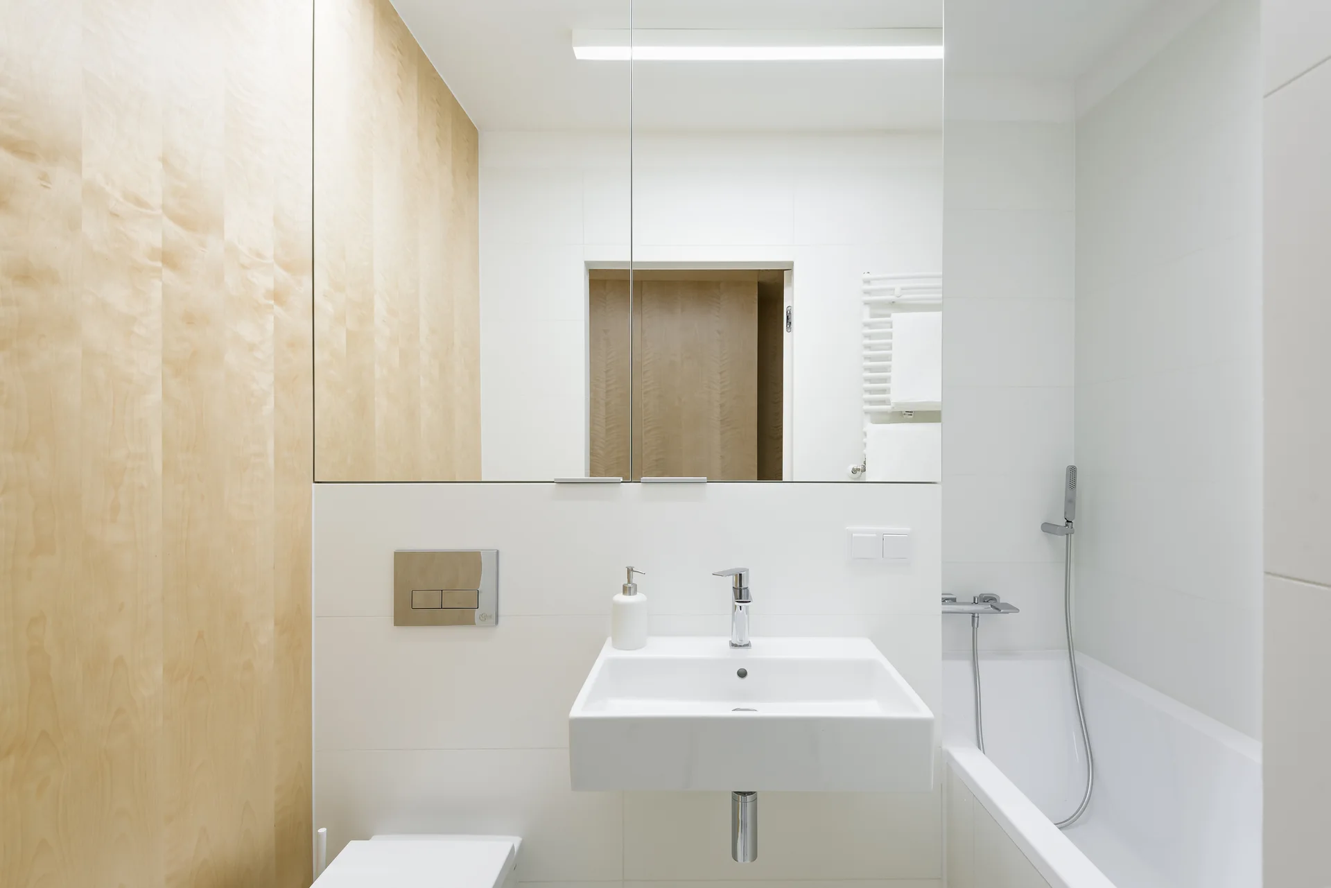A minimalist white bathroom features light wood wall paneling, a large rectangular wall mirror, a wall-mounted sink, and a white bathtub.