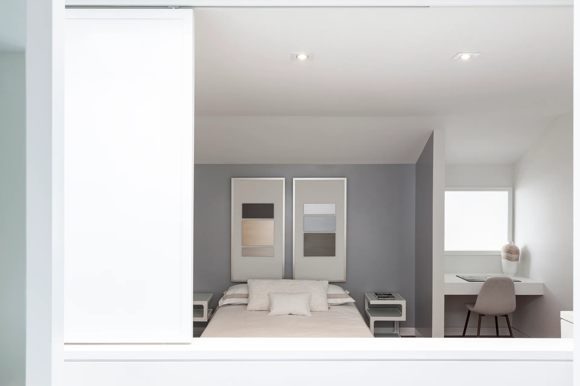 A minimalist bedroom featuring a bed flanked by white bedside tables, a gray accent wall, and a built-in white desk inside a side alcove.