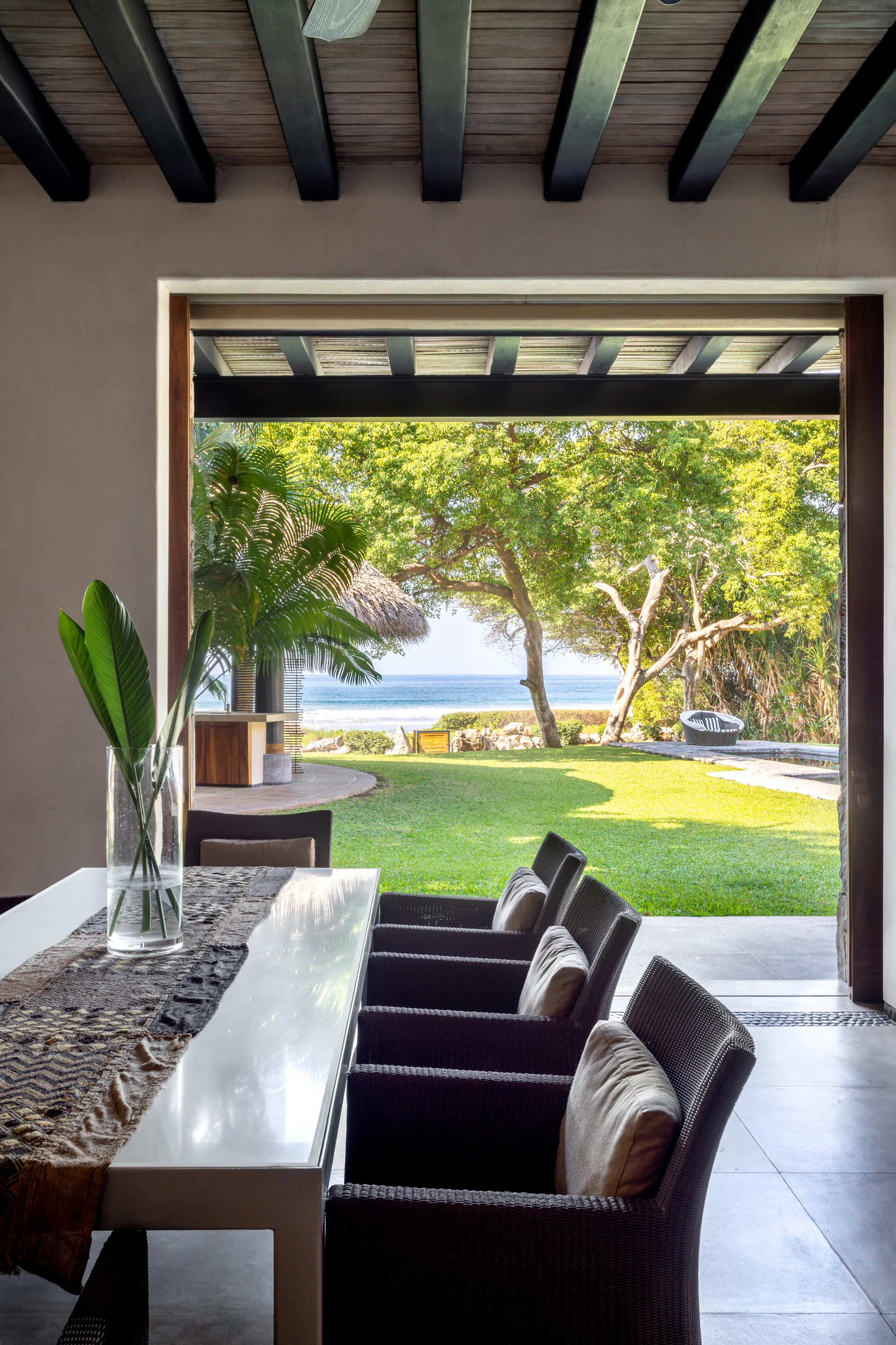 An indoor dining table with woven chairs sits under dark wooden ceiling beams, looking out an open wall to a grassy lawn and the ocean.