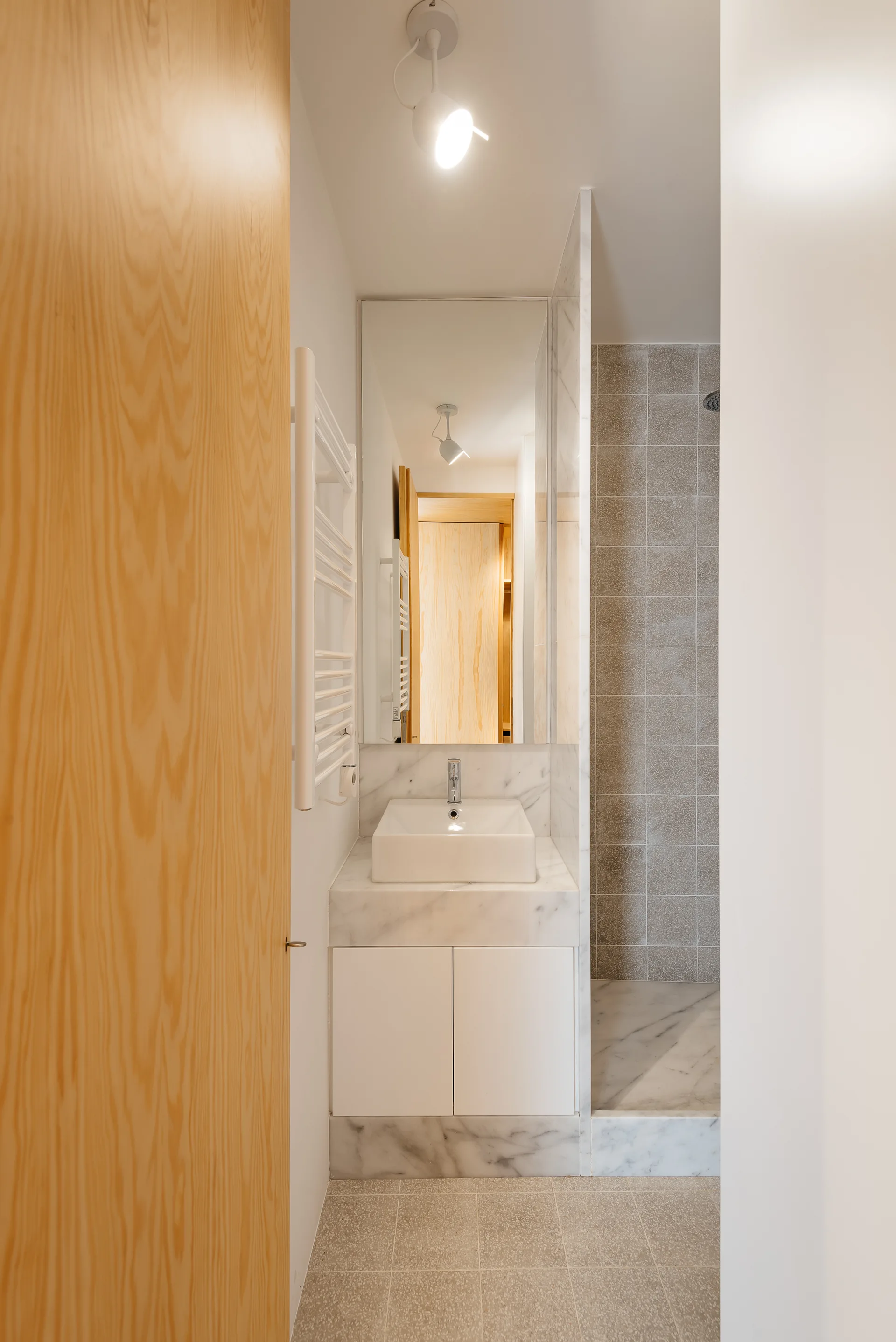 A narrow modern bathroom features a white marble vanity with a vessel sink, a tall rectangular mirror, and a tiled walk-in shower.