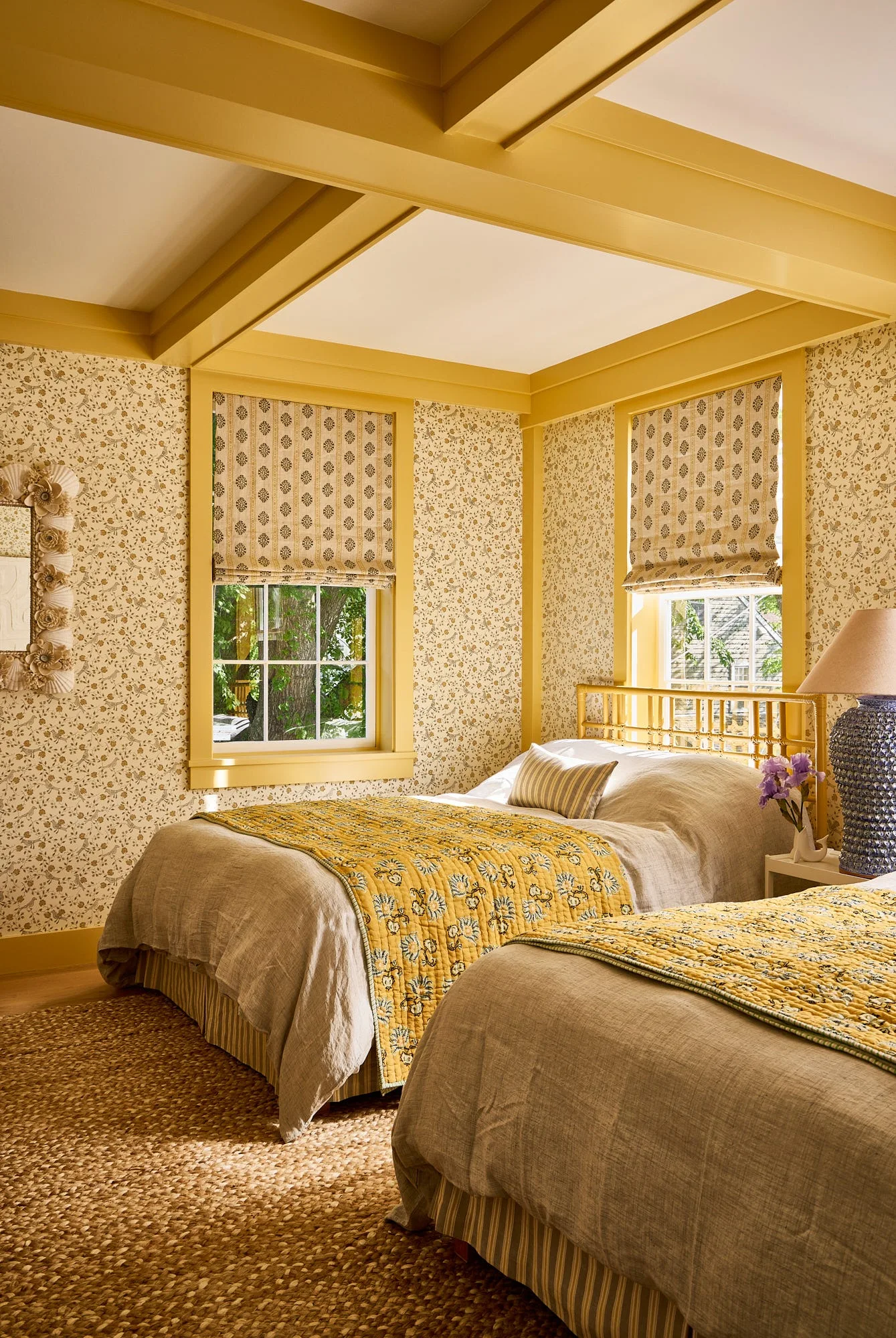 Two twin beds with yellow quilted covers sit in a bedroom featuring floral wallpaper, yellow-painted ceiling beams, and matching window trim.