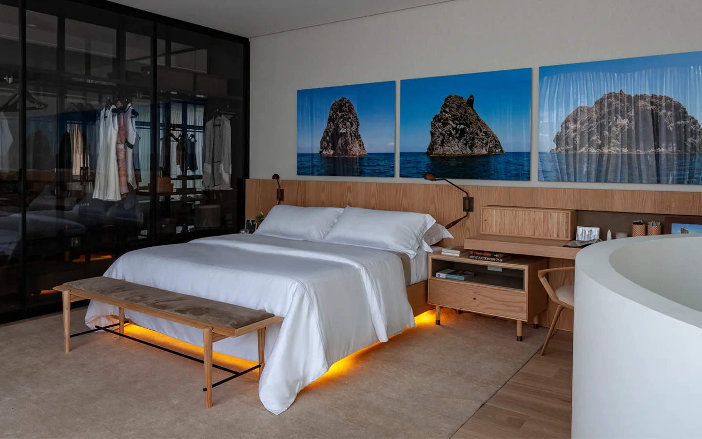 A modern bedroom features a bed with white linens, an integrated light wood headboard and desk system, and a black-framed glass closet.