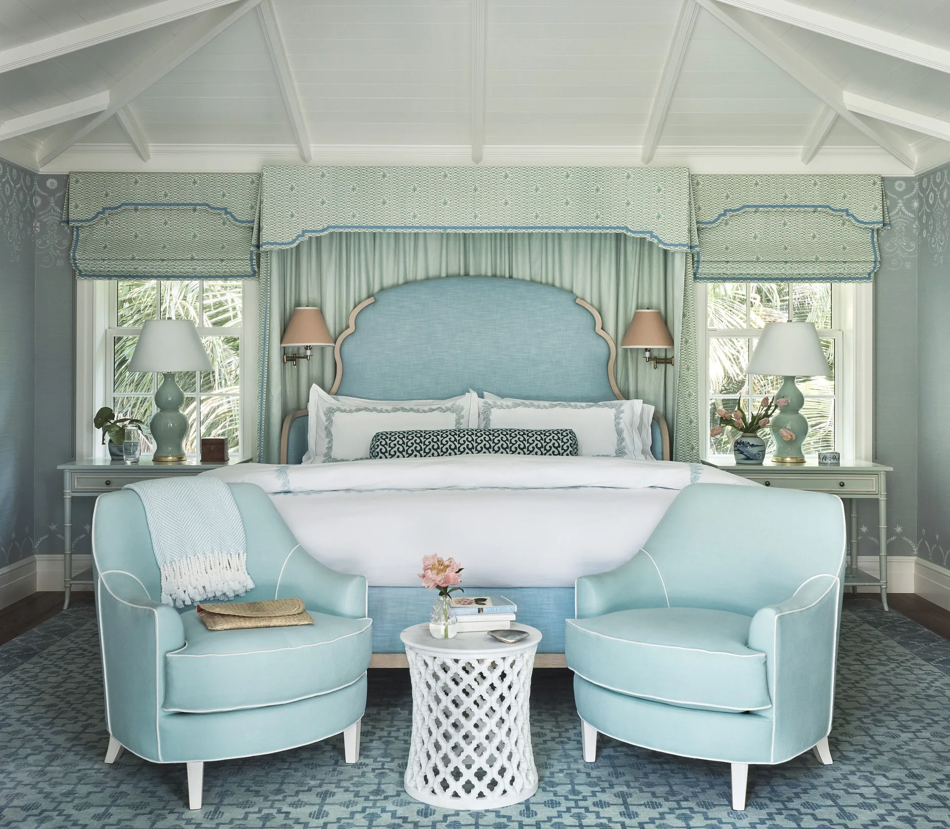 A light blue bedroom featuring a canopy bed, two matching armchairs with white piping, a geometric rug, and a vaulted beamed ceiling.