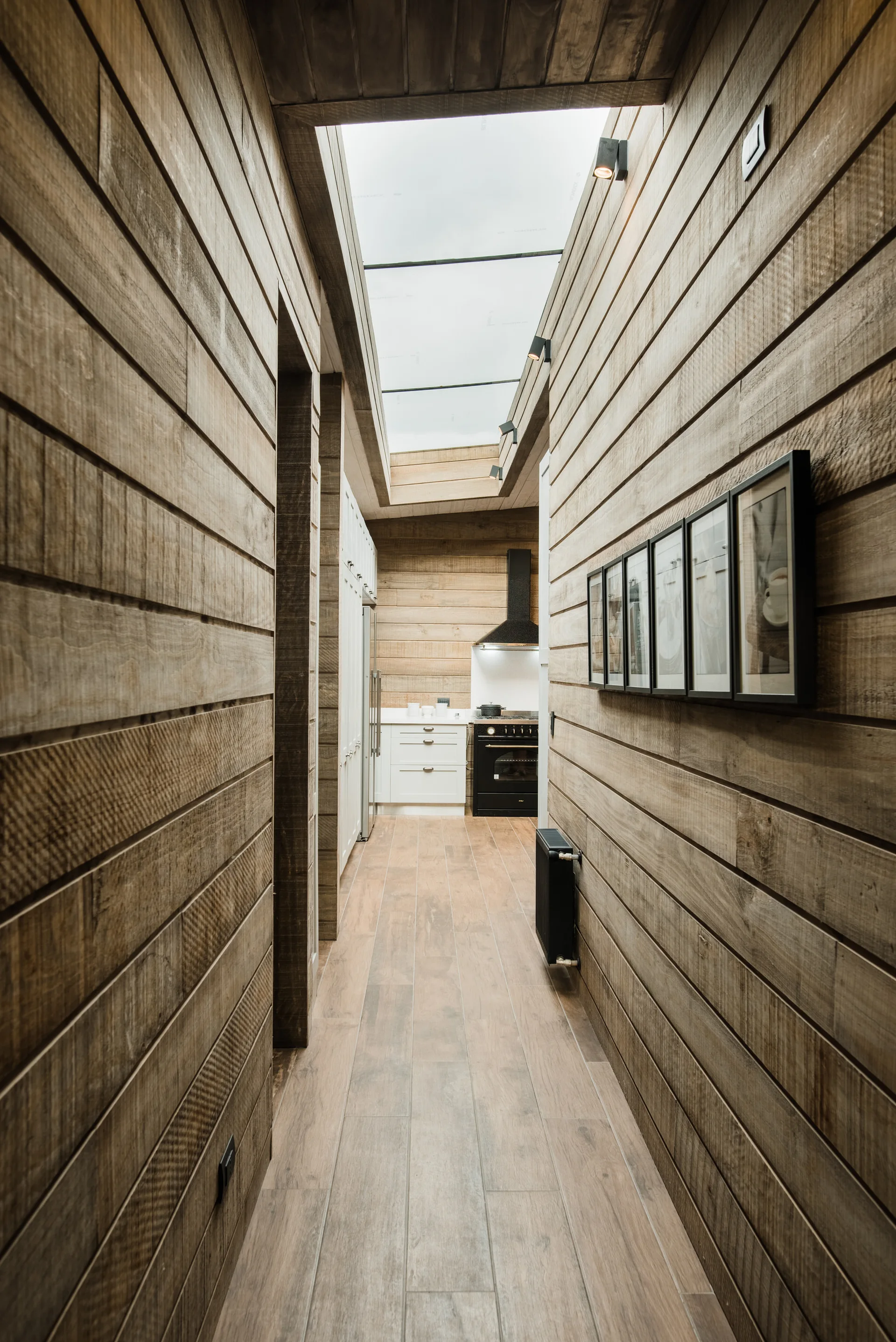A narrow hallway with horizontal wood plank walls and an overhead skylight leading toward a white kitchen in the background.