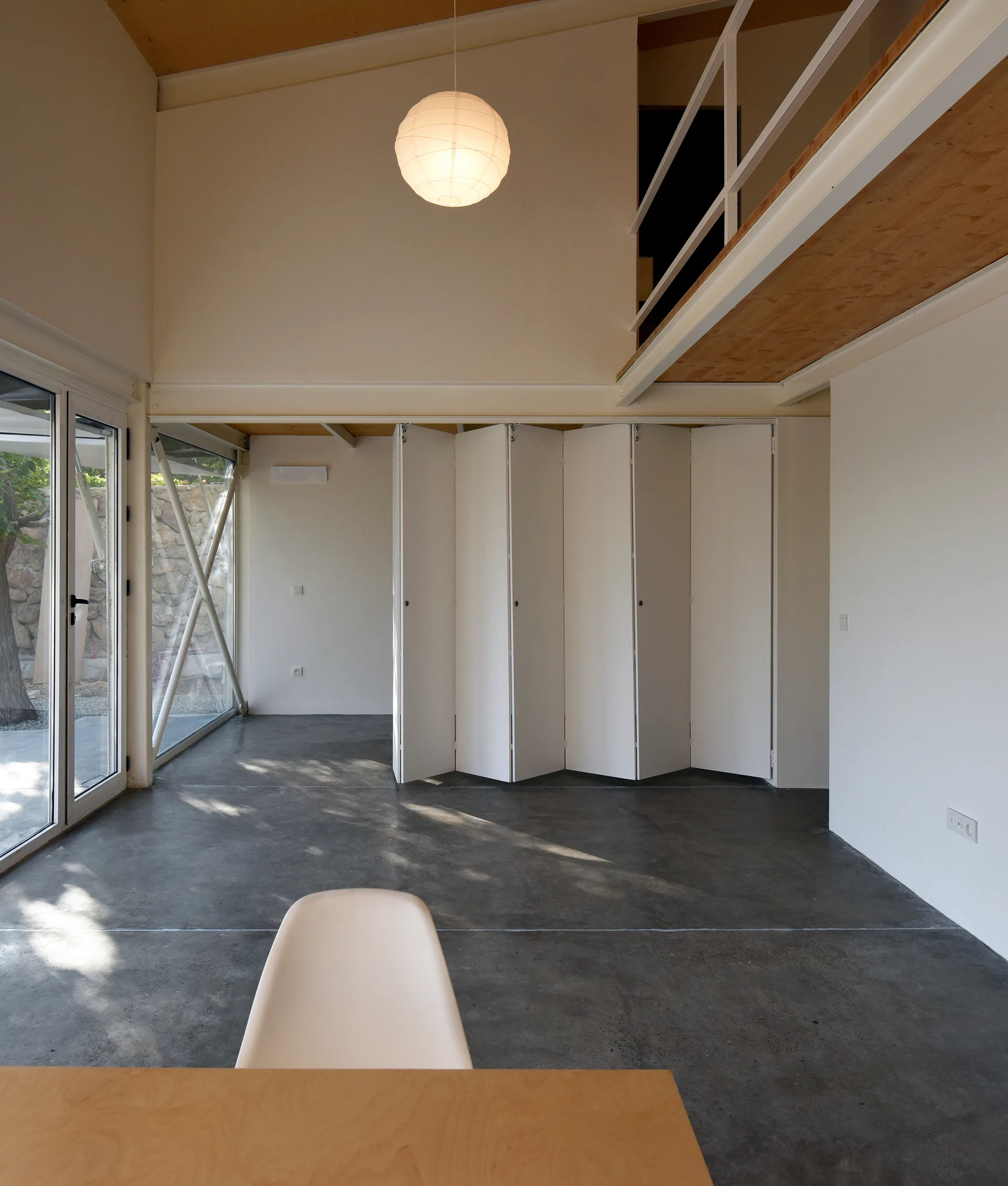 A minimalist loft space featuring dark polished concrete floors, a large white folding partition wall, and a mezzanine balcony with wood ceilings.