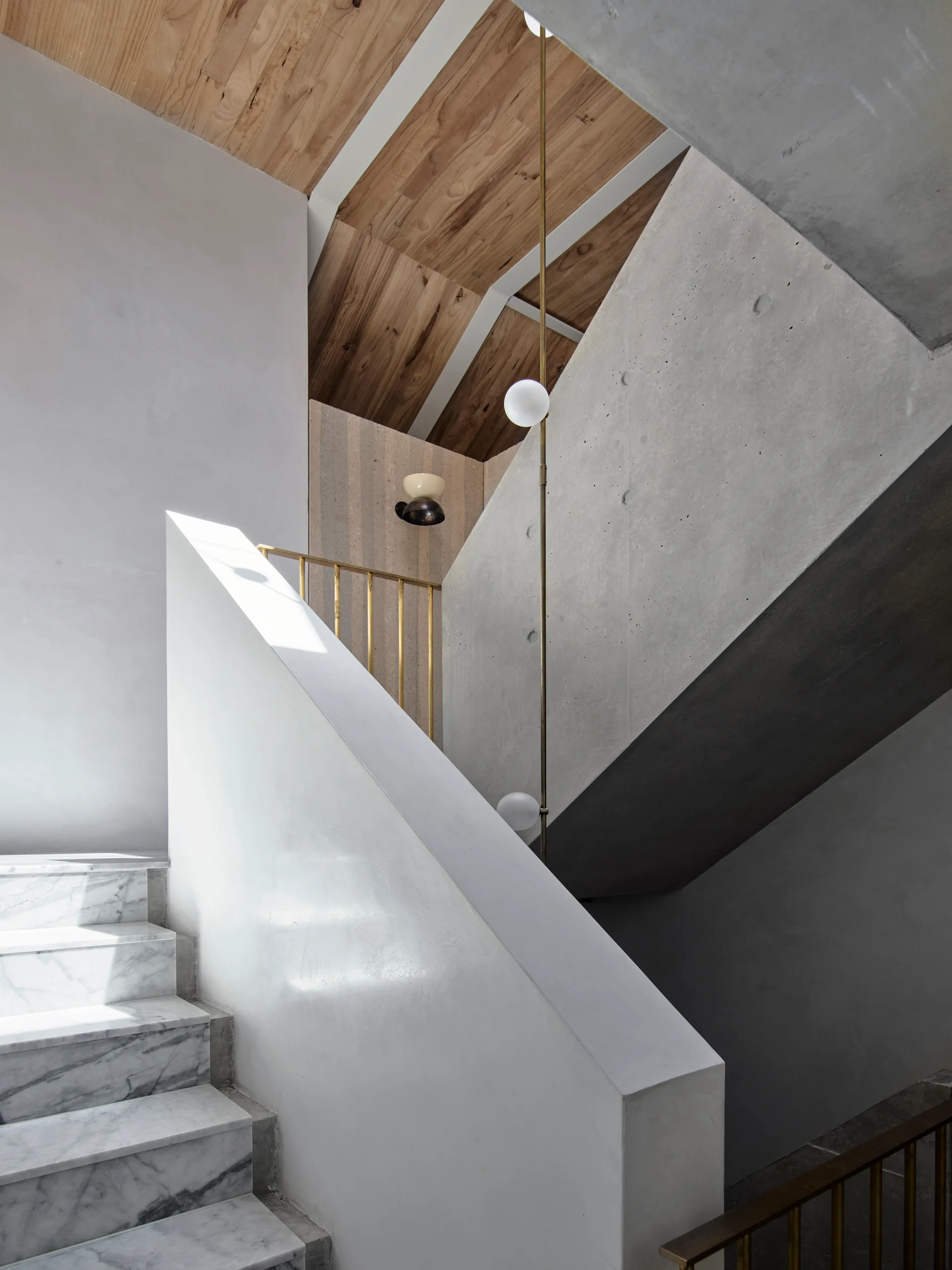 A modern staircase features white veined marble steps, a raw grey concrete wall, vertical brass railings, and a wood plank ceiling.
