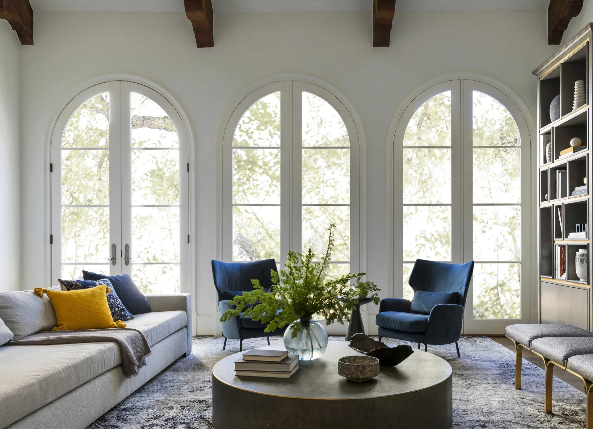 A bright living room with three large arched glass doors, a grey sectional sofa, two blue armchairs, and a round metal coffee table on a patterned rug.