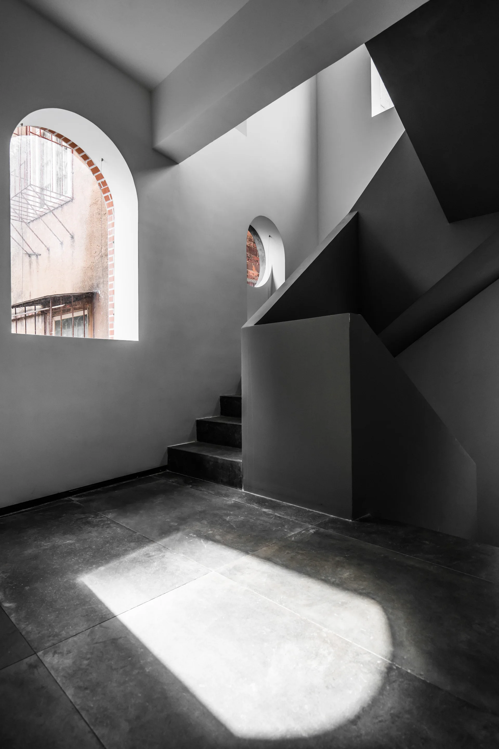 A minimalist gray plaster staircase next to arched windows that look out onto a brick facade.