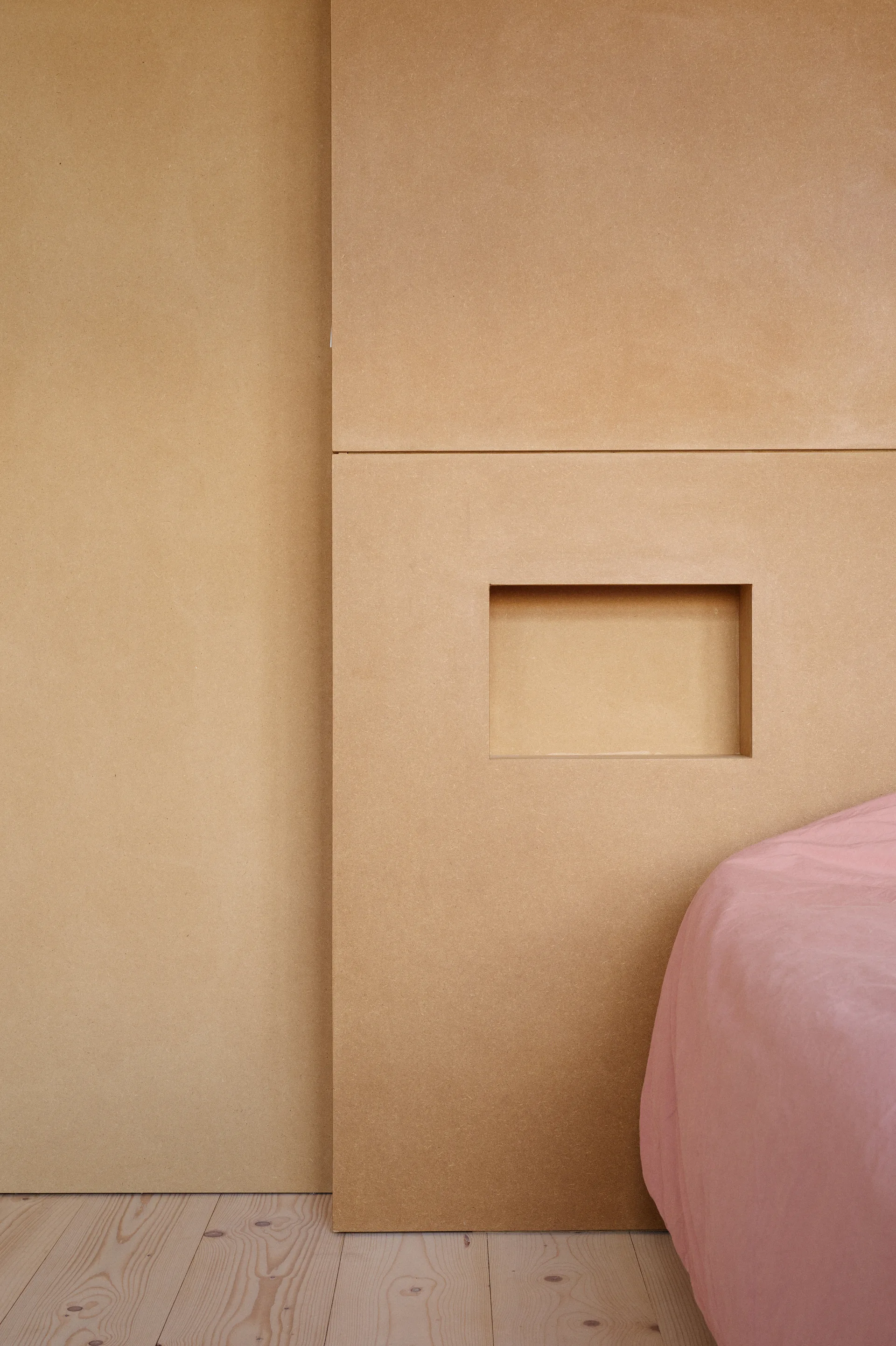 Minimalist tan MDF wall panels with a small rectangular recessed niche next to a bed with dusty pink bedding.