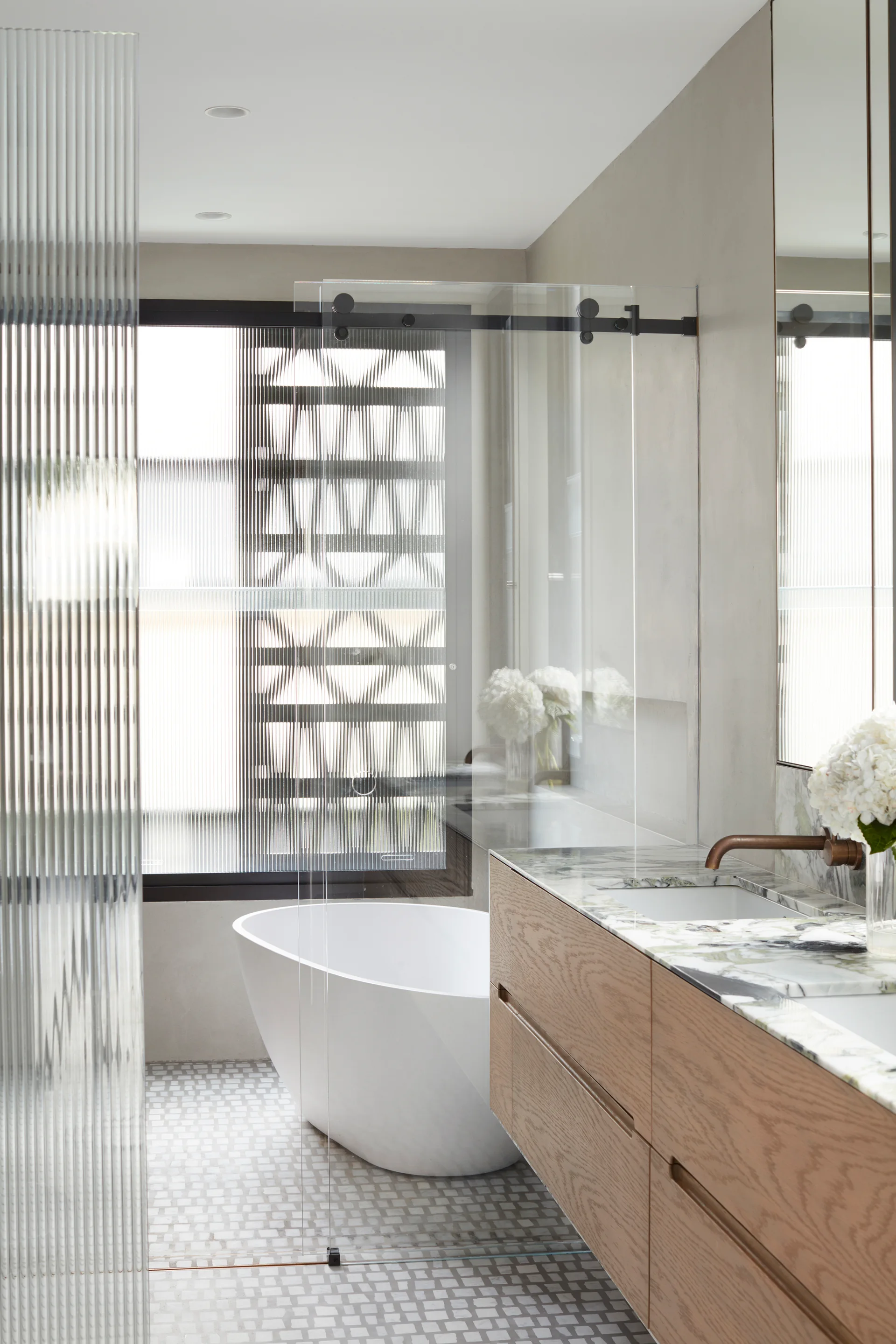 A modern bathroom features a light wood vanity with a marble countertop, a white freestanding tub, and a fluted glass partition.