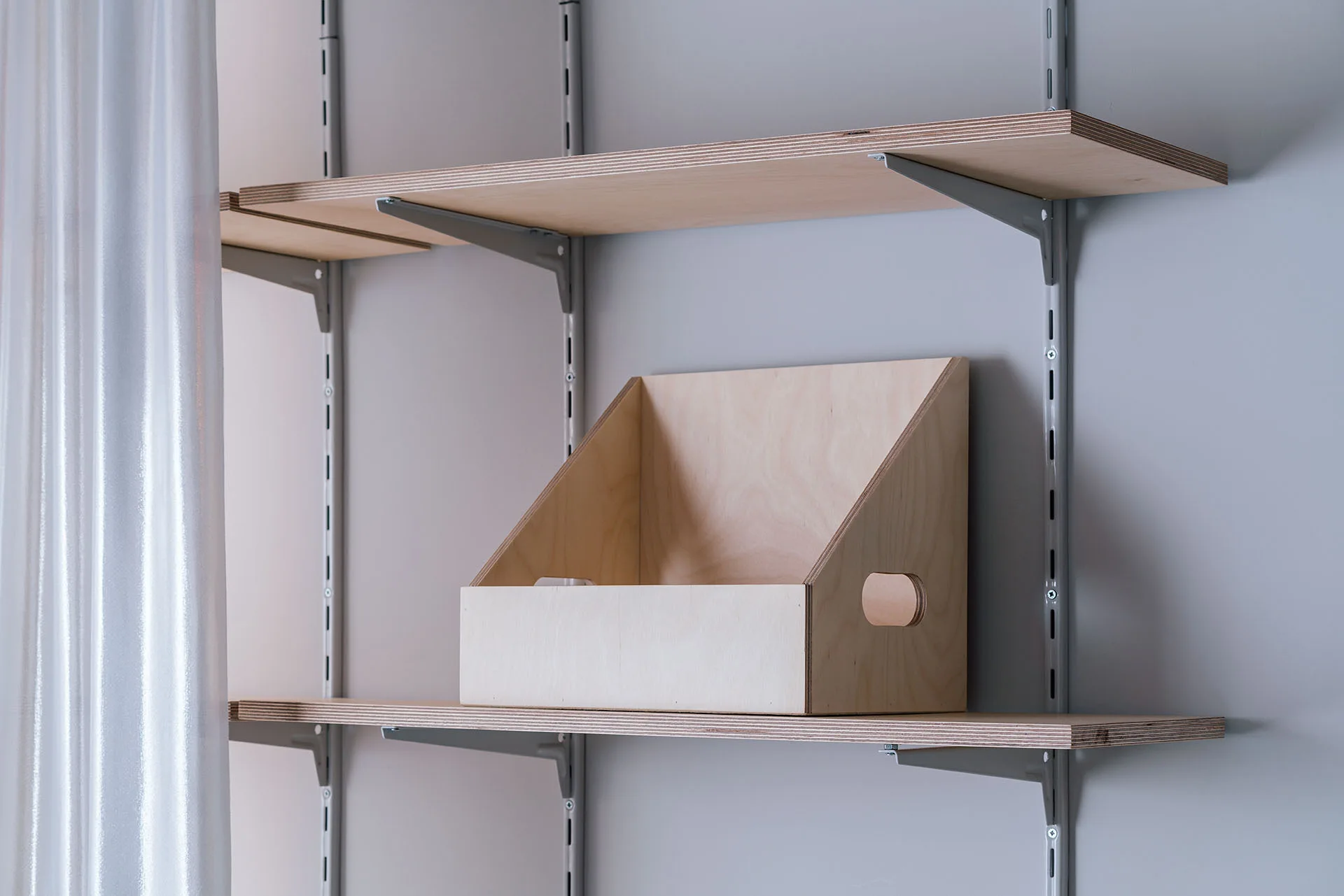 A wall-mounted plywood shelving system with adjustable metal tracks holding a matching wooden storage box.