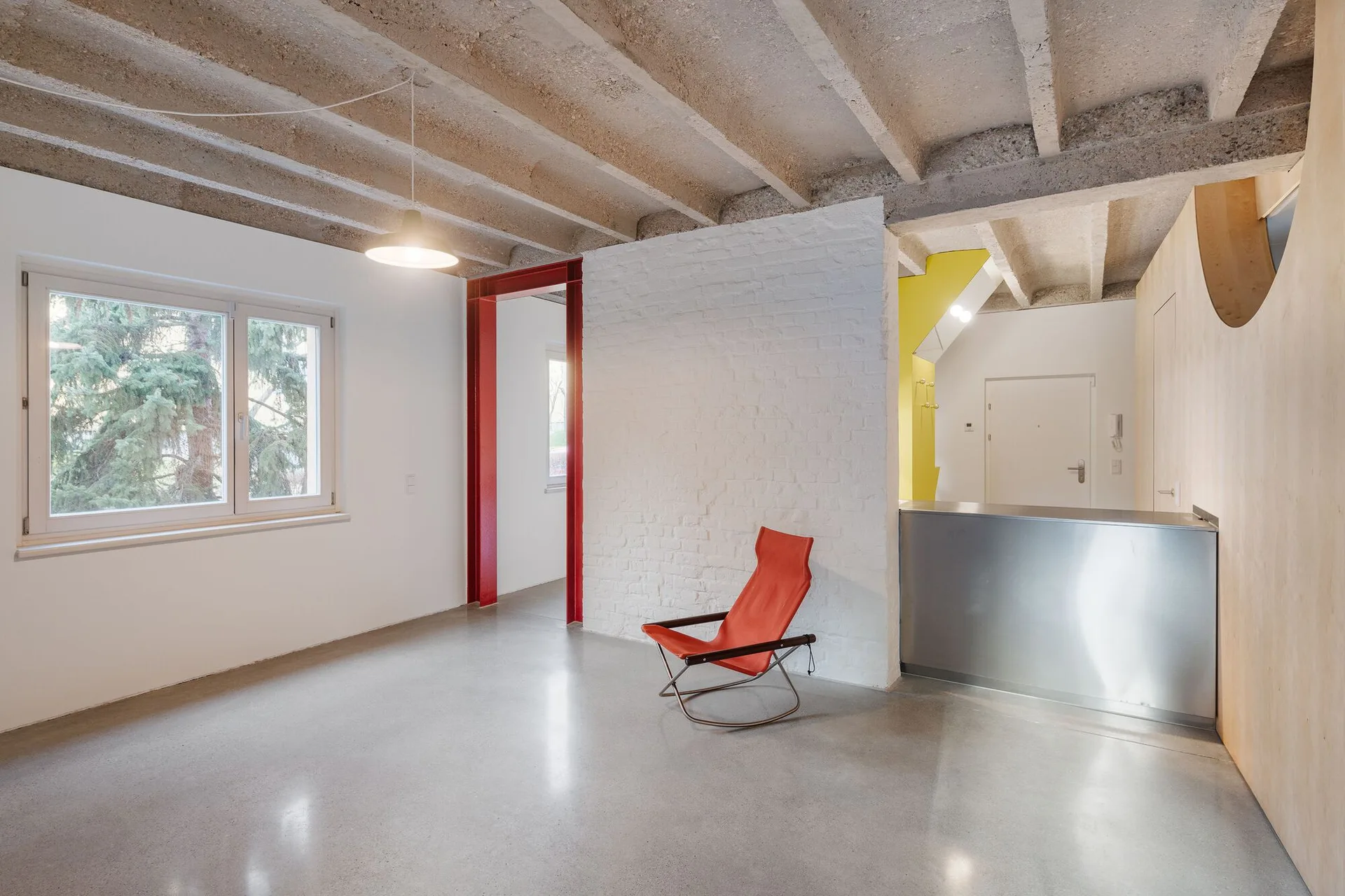 A minimalist room featuring an exposed concrete beam ceiling, polished concrete floor, painted white brick wall, and a bright red doorframe.