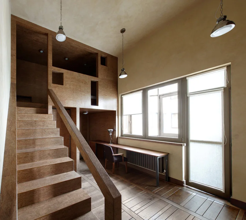 A wooden staircase leads to a loft platform next to a long desk built under a large window in a room with plaster walls.