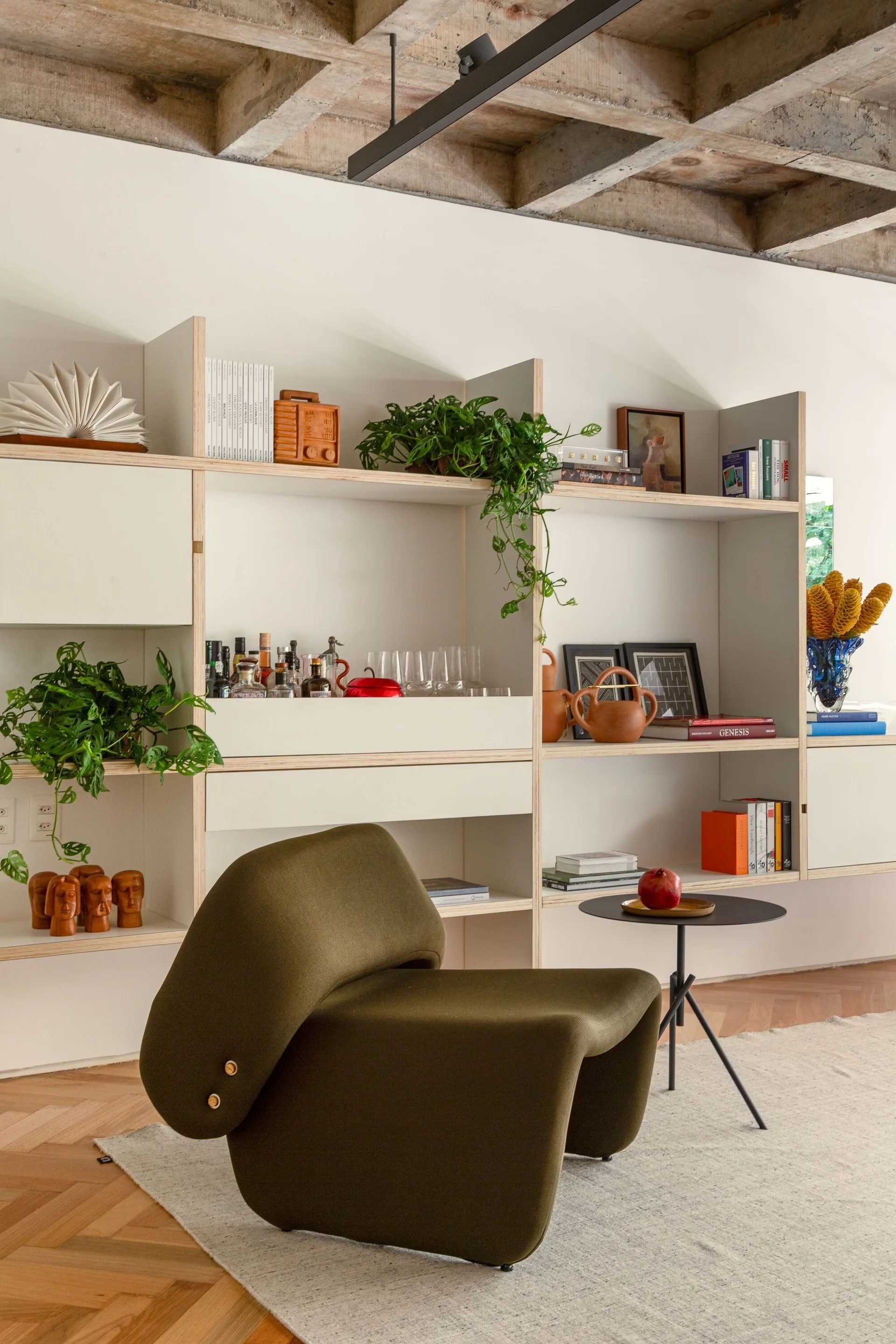 An olive green sculptural lounge chair sits in front of a large plywood shelving unit filled with books and plants under a raw concrete ceiling.