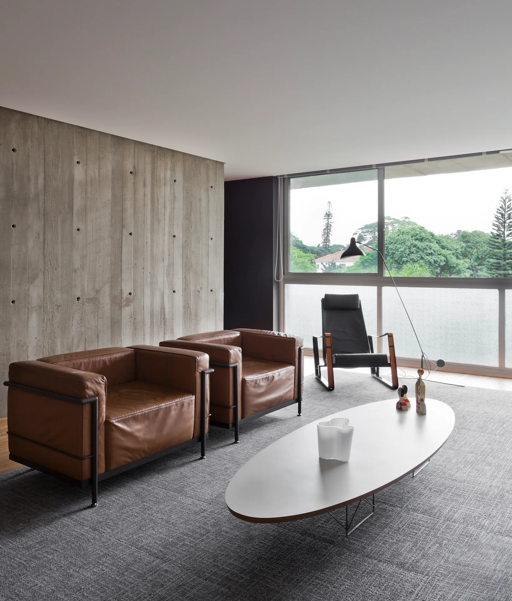 A minimalist living room features two brown leather armchairs, an oval white coffee table, a textured concrete wall, and a large window.
