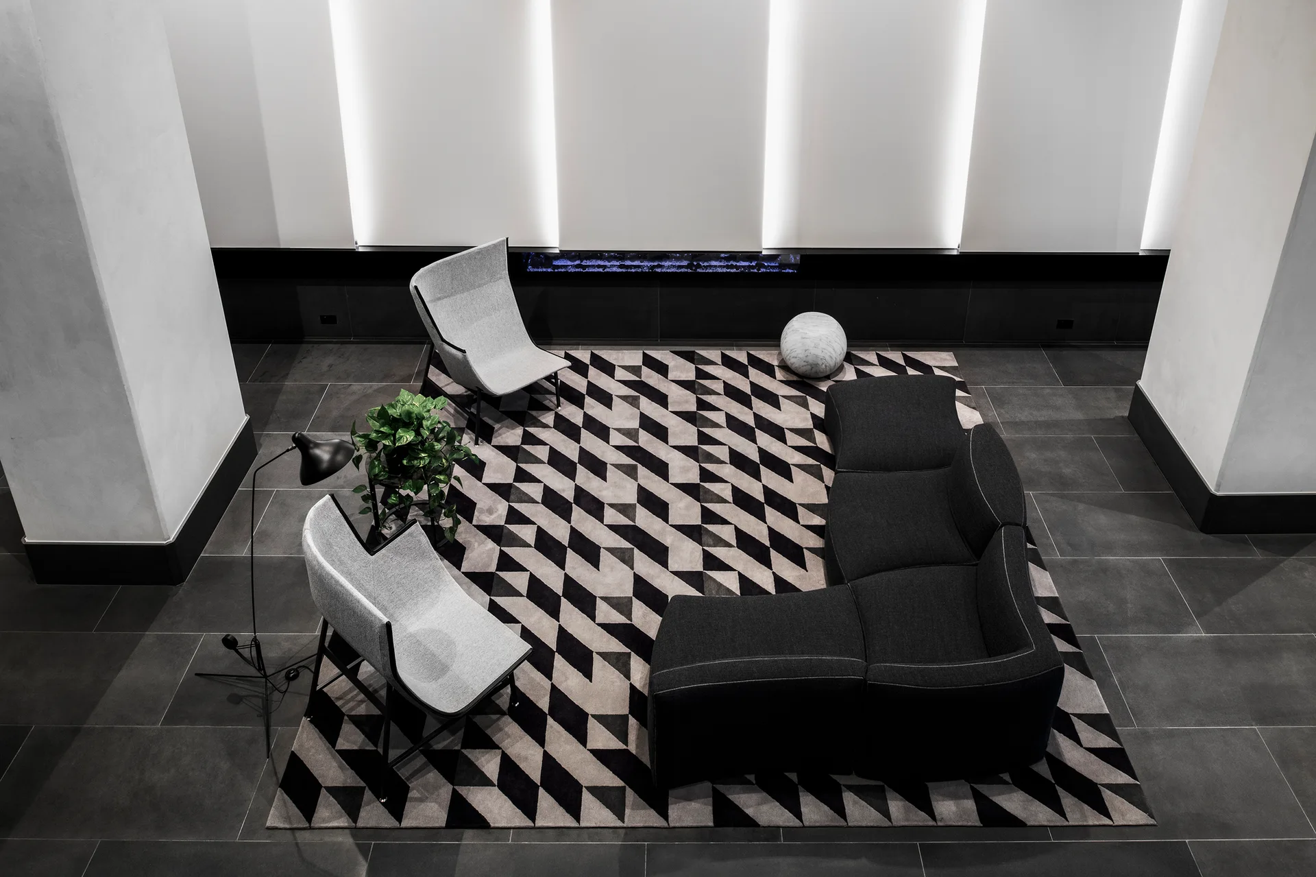 A high-angle view of a modern lounge featuring a dark modular sofa, two light grey chairs, and a bold geometric-patterned rug.