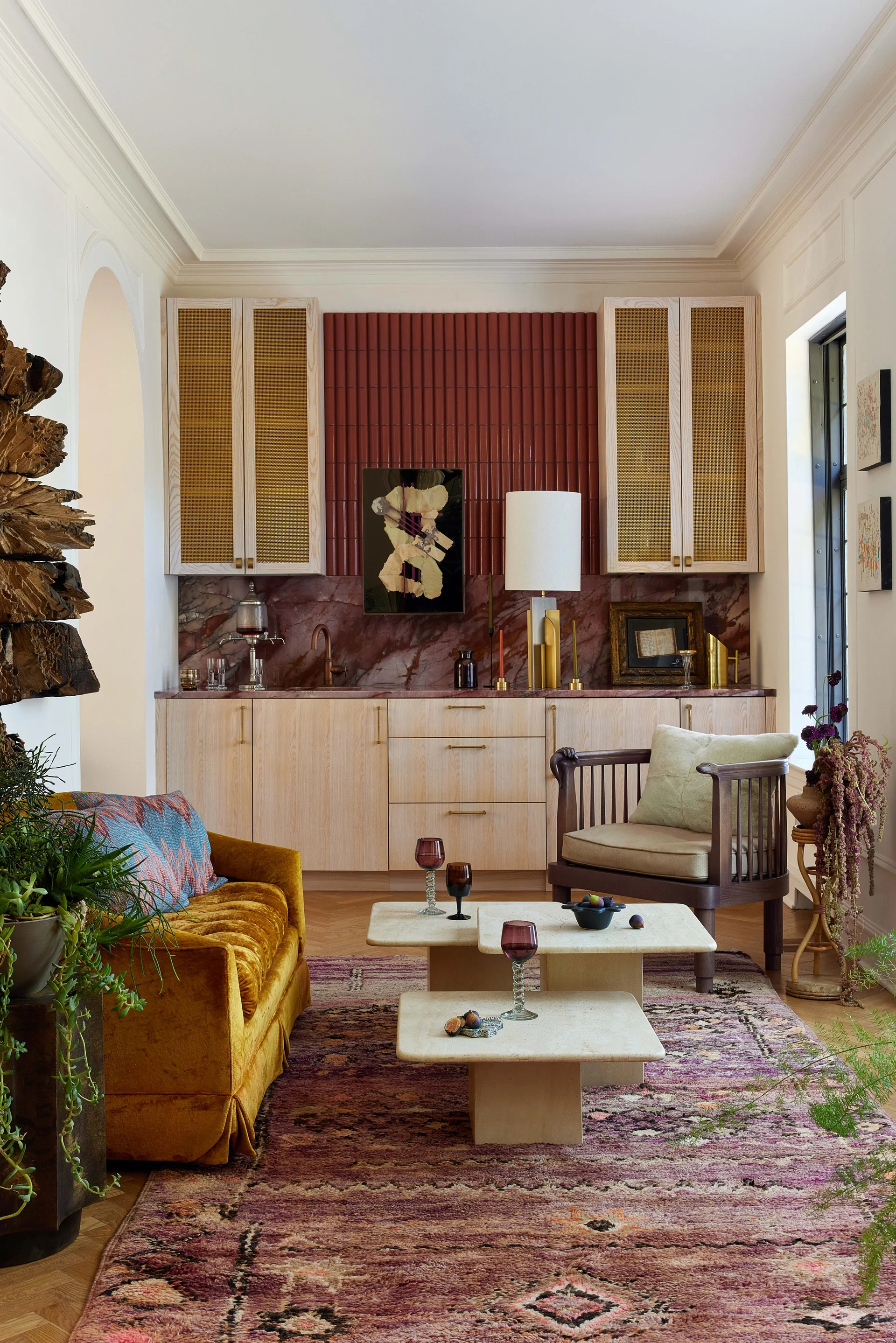 A lounge features a built-in wooden bar with a red tile backsplash, a mustard velvet sofa, and stacked stone coffee tables on a patterned rug.