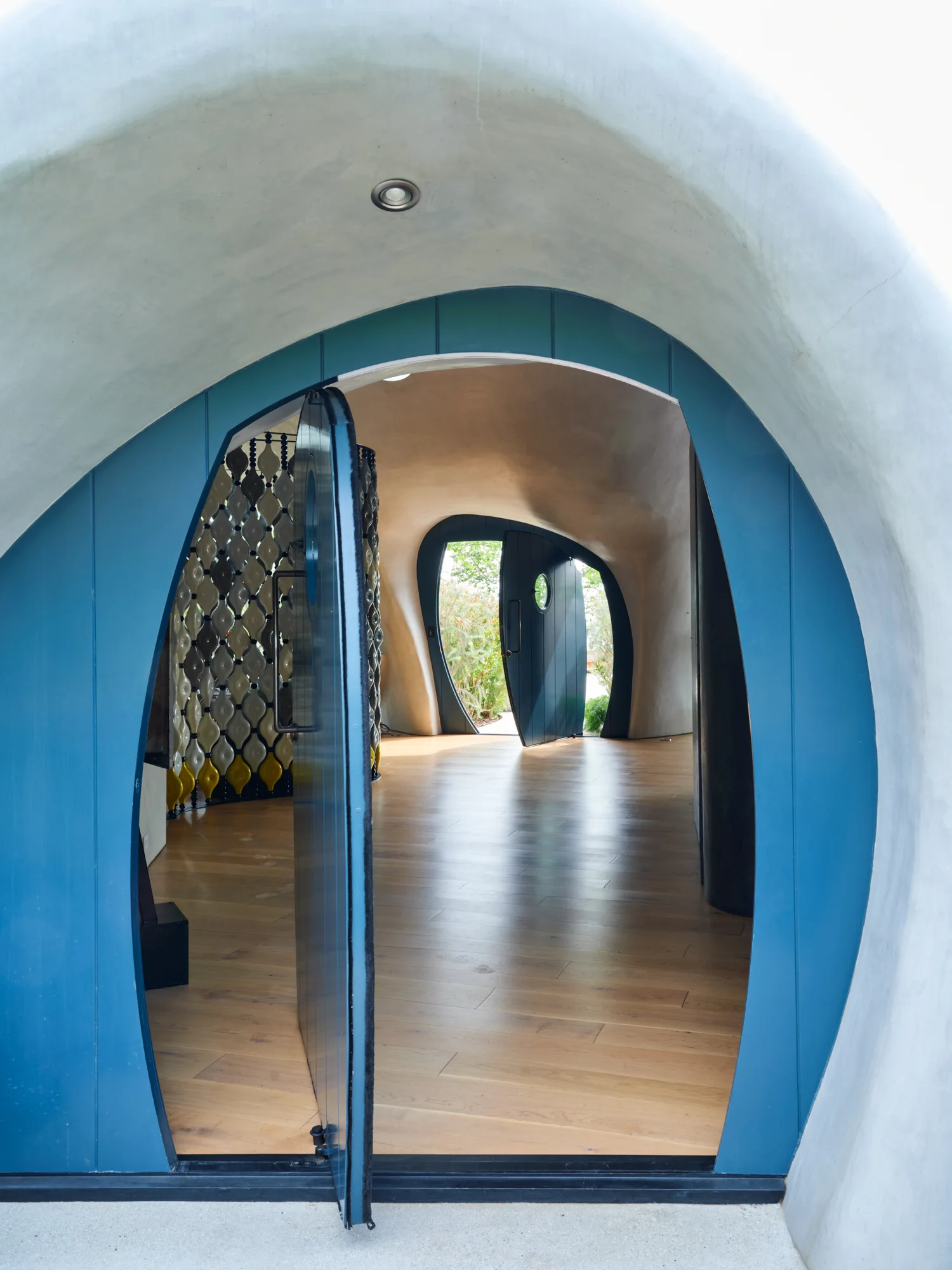 An organic entryway features curved grey plaster walls, light wood flooring, and distinct oval-shaped blue doors that frame the path.