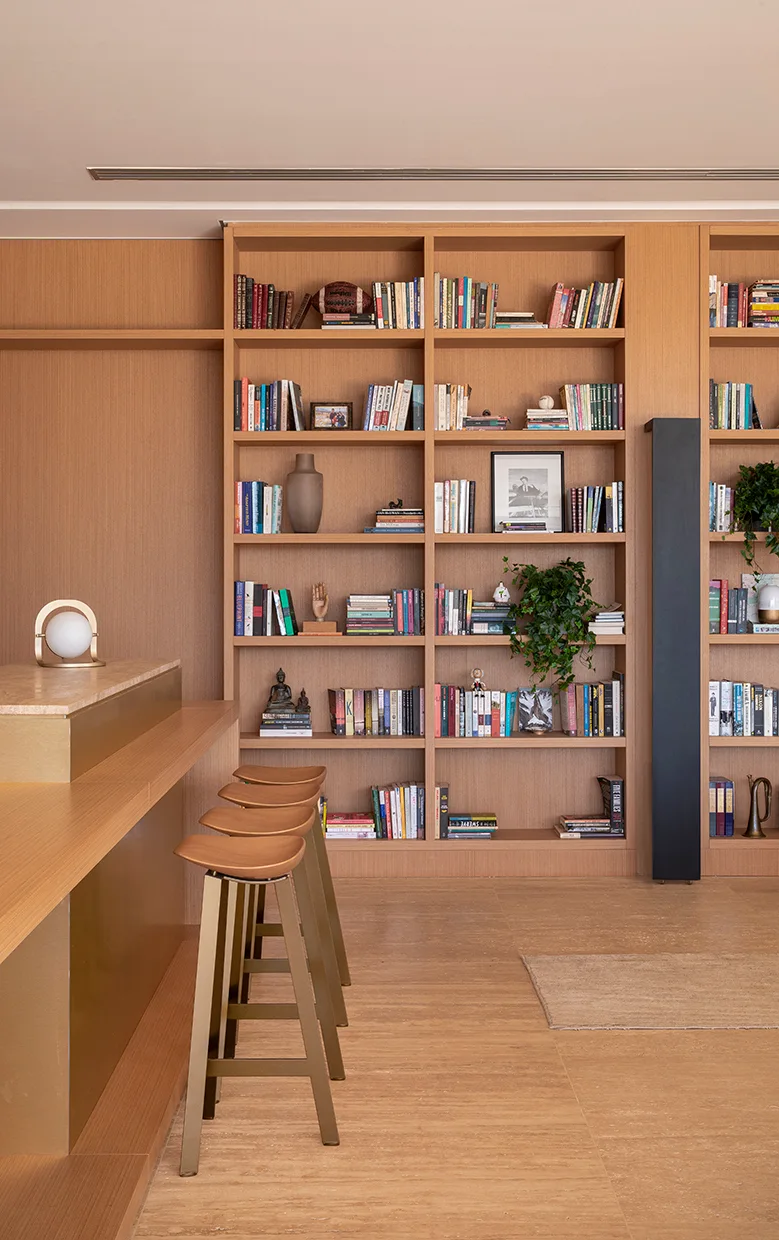 A floor-to-ceiling wooden bookshelf filled with books sits behind a light wood bar counter with three curved wooden bar stools.