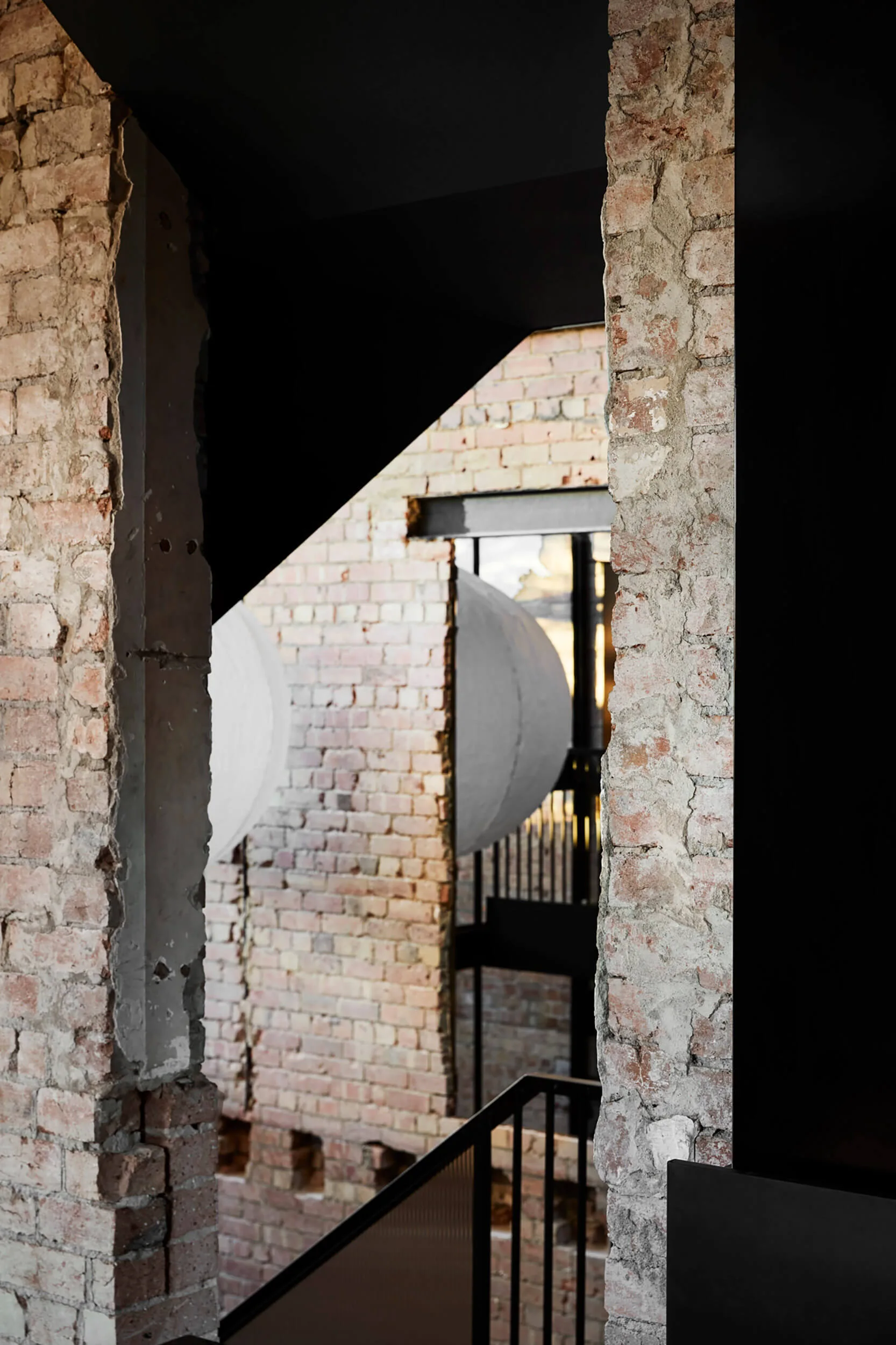 A modern stairwell features rough exposed brick walls with jagged cutouts, a black metal railing, and a large white sphere in the background.