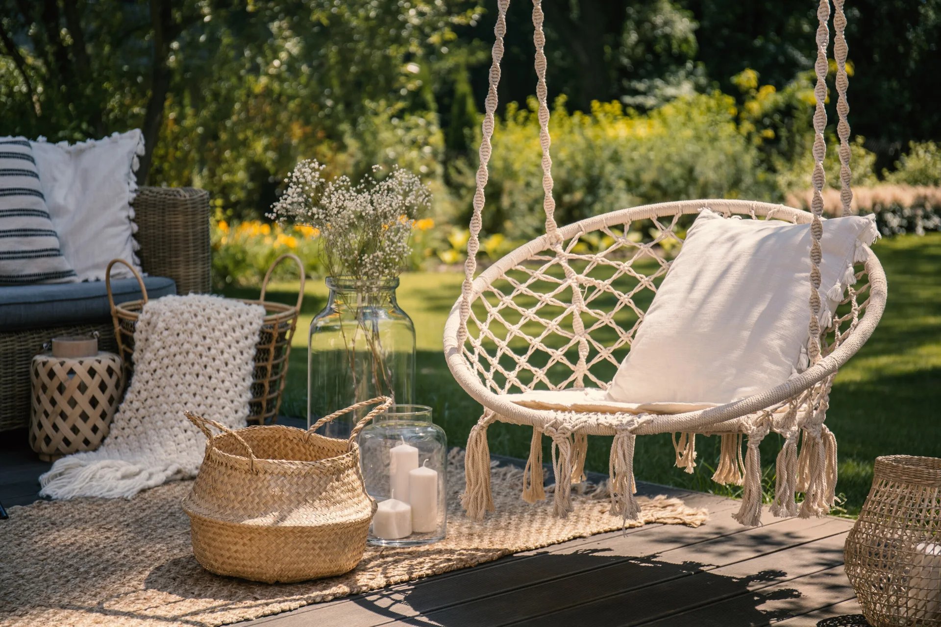 A wooden patio deck features a hanging macrame swing chair with a white cushion, surrounded by woven baskets, glass candle holders, and a natural fiber rug.