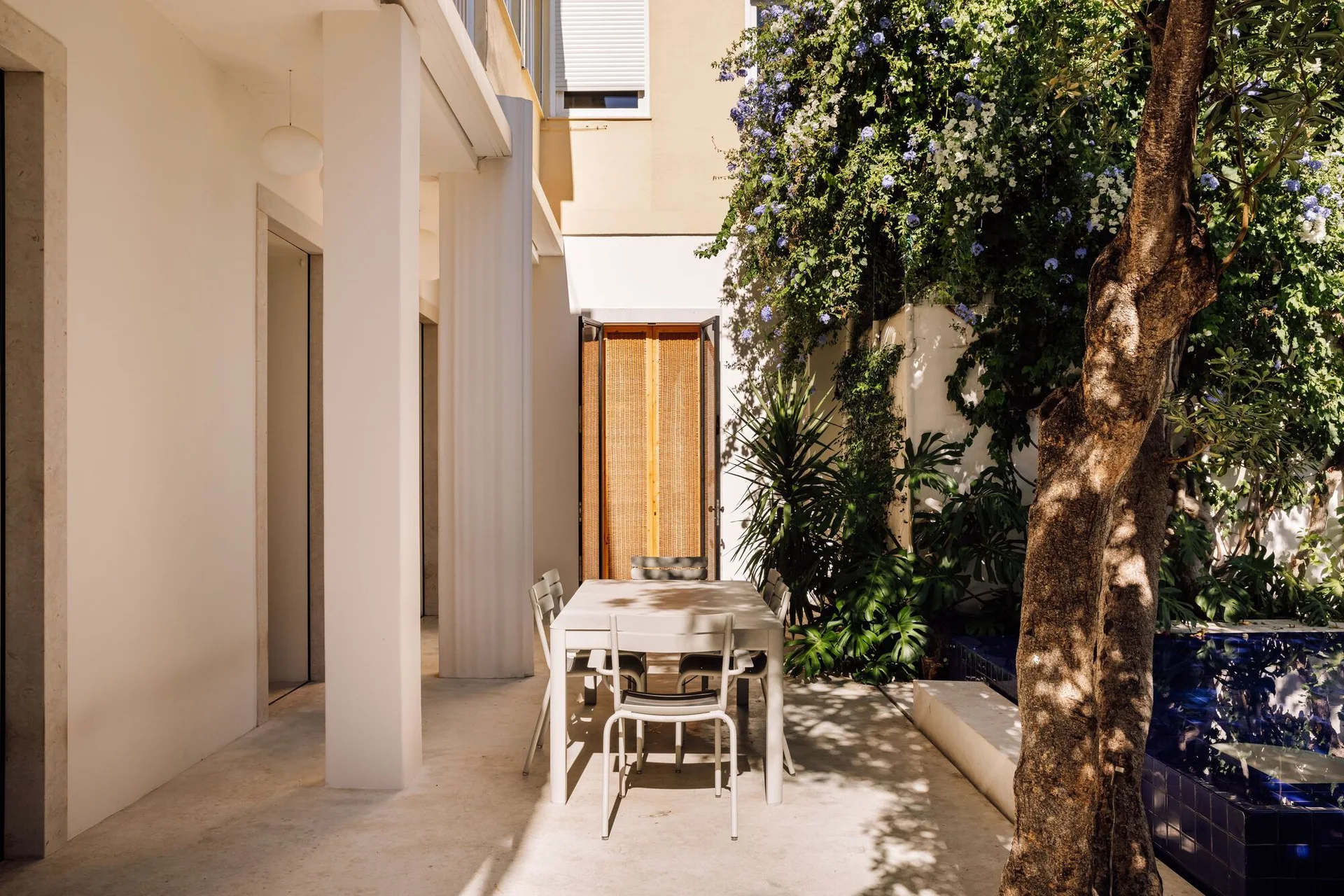 A white outdoor dining set sits on a concrete patio next to a blue tiled water feature, bordered by white columns and lush climbing vines.