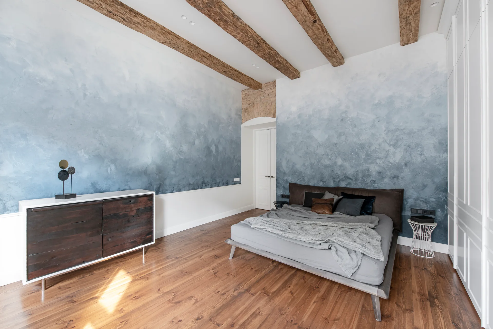 A bedroom featuring a textured blue and white ombre wall, natural wood ceiling beams, a low grey bed, and hardwood flooring.