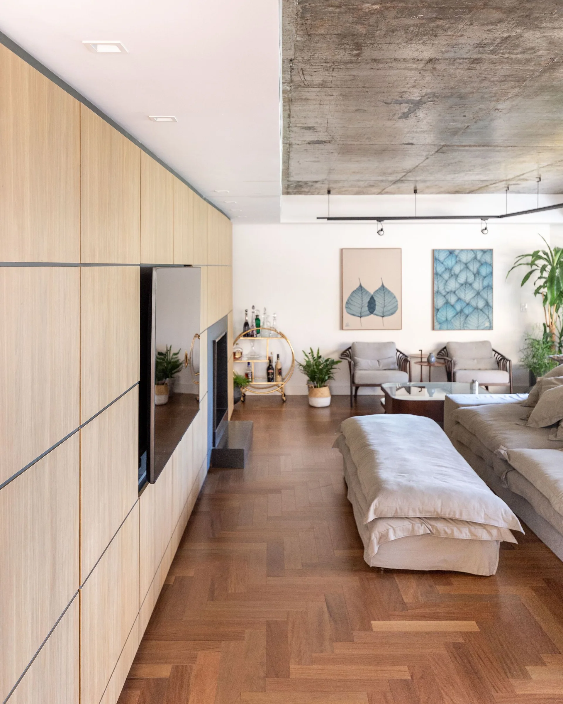 A modern living room features a floor-to-ceiling wood paneled wall with an integrated television, herringbone wood floors, and a raw concrete ceiling.