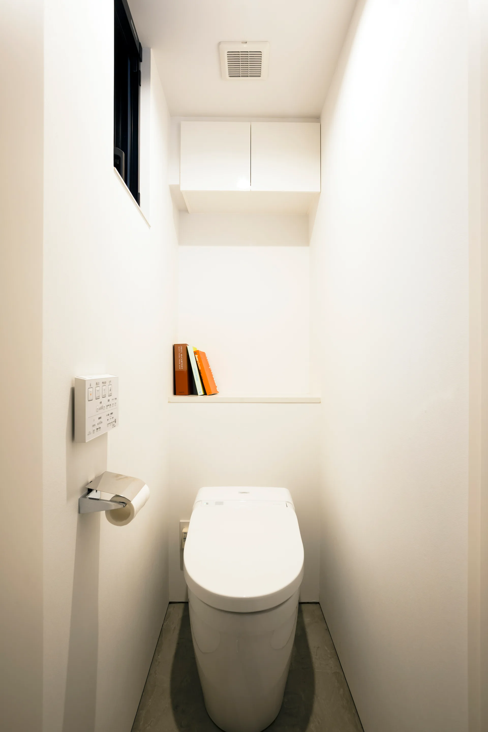 A minimalist all-white powder room featuring a modern toilet, grey concrete floor, a recessed shelf holding books, and a high storage cabinet.