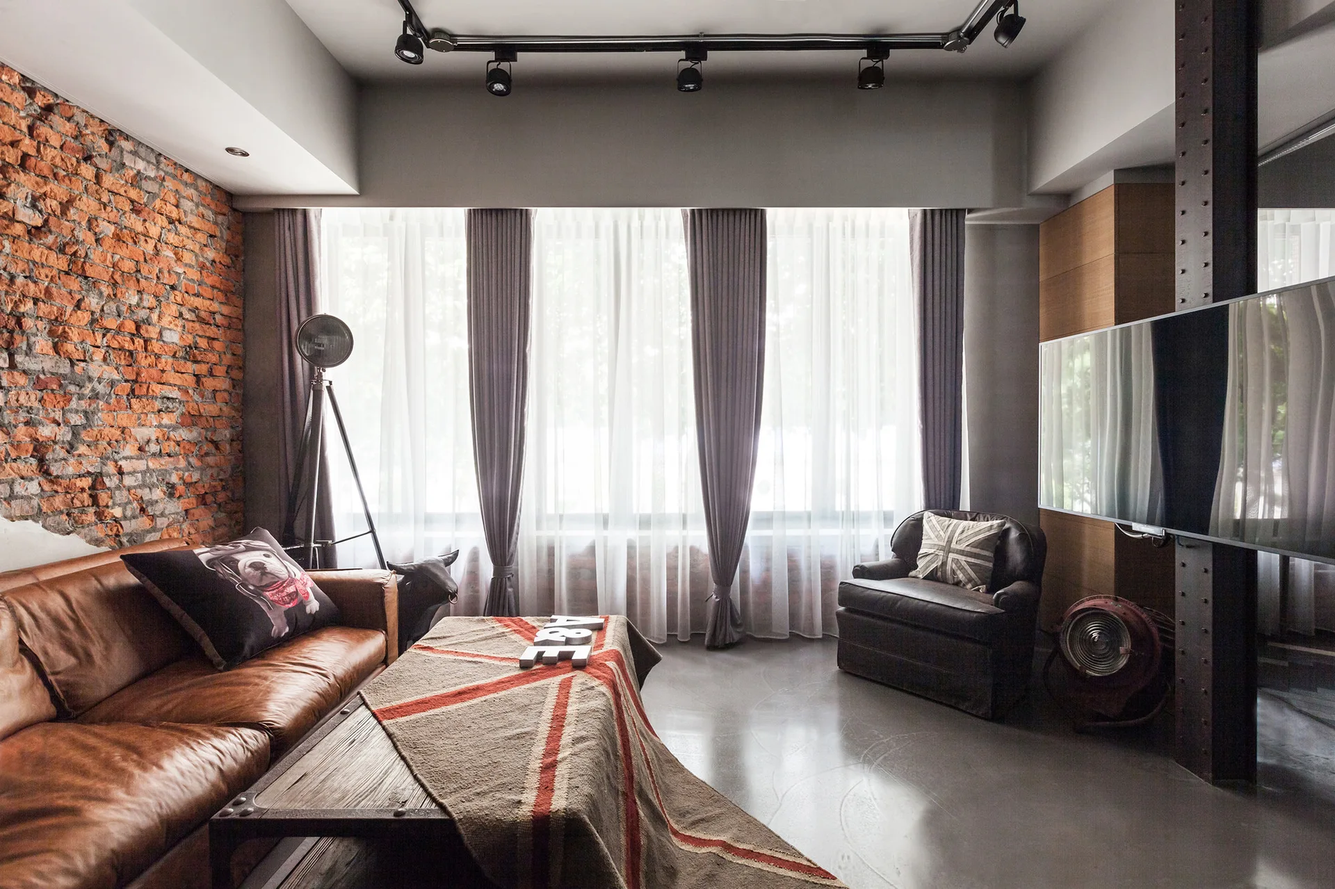An industrial loft living room with an exposed brick wall, a brown leather sofa, and large windows covered by sheer curtains.
