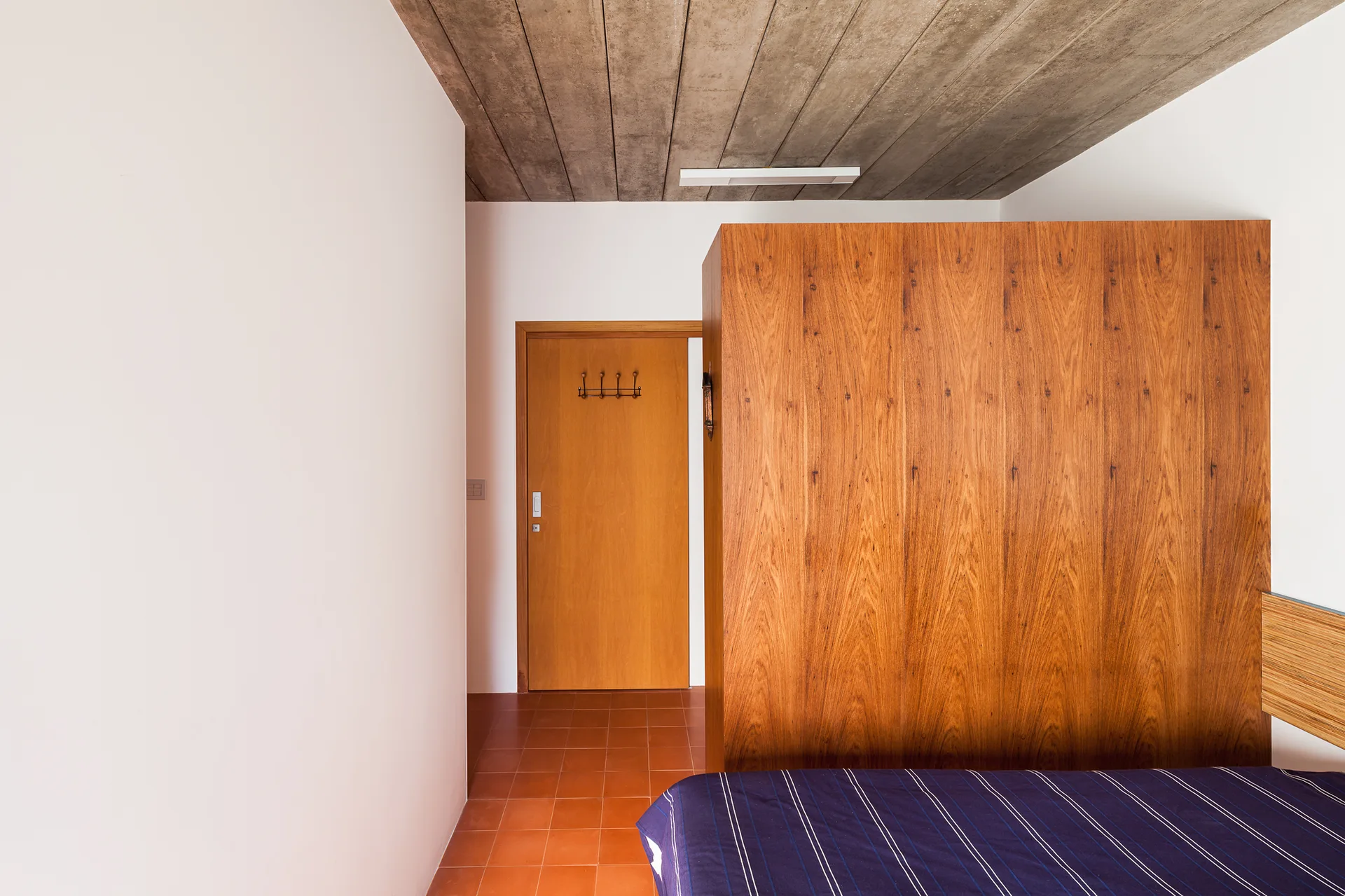 A bedroom featuring a large wood grain partition wall, terracotta floor tiles, and a textured concrete ceiling above a striped bed.