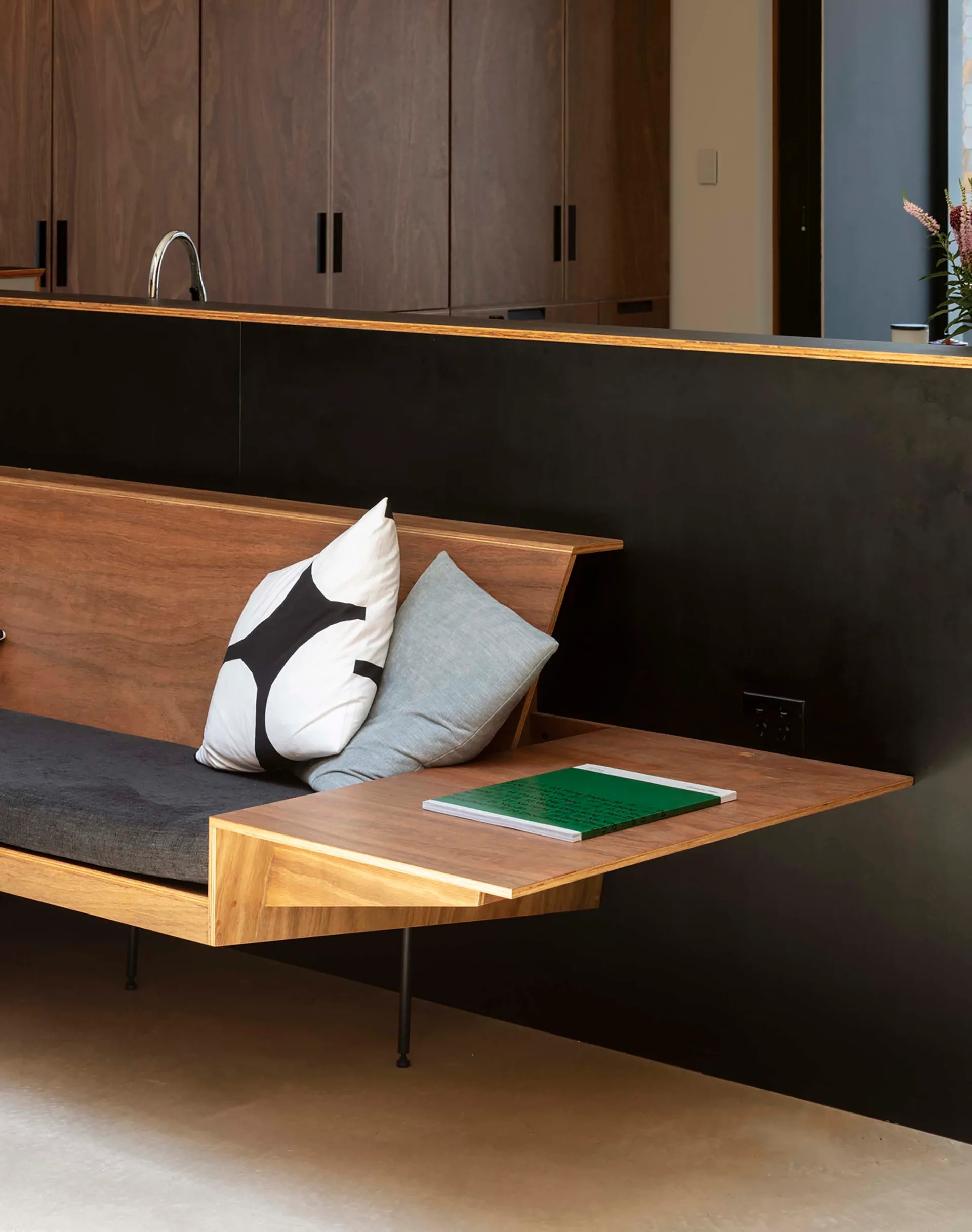 A modern wood sofa with black and grey cushions is integrated into a black partition wall, featuring a cantilevered side table extension.