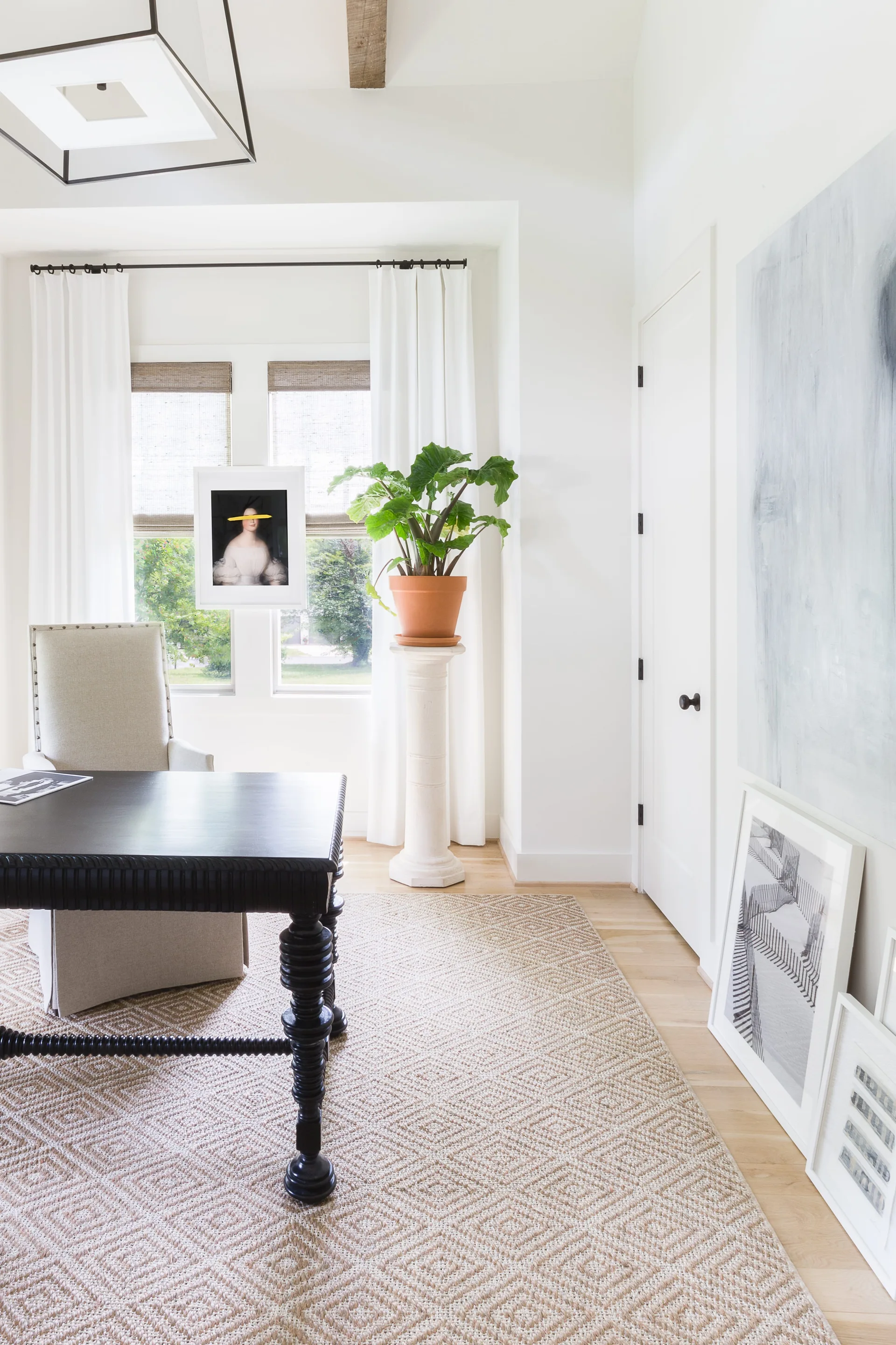 A bright home office features a dark wooden desk, a light beige upholstered chair, and a green plant sitting on a tall stone column pedestal.