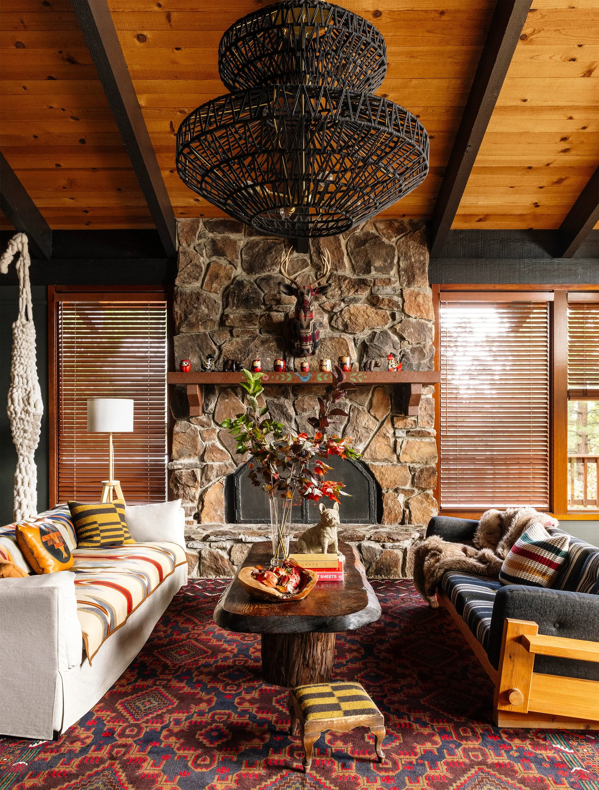 A rustic living room featuring a floor-to-ceiling stone fireplace, a wood paneled ceiling with dark beams, and a large black woven chandelier.