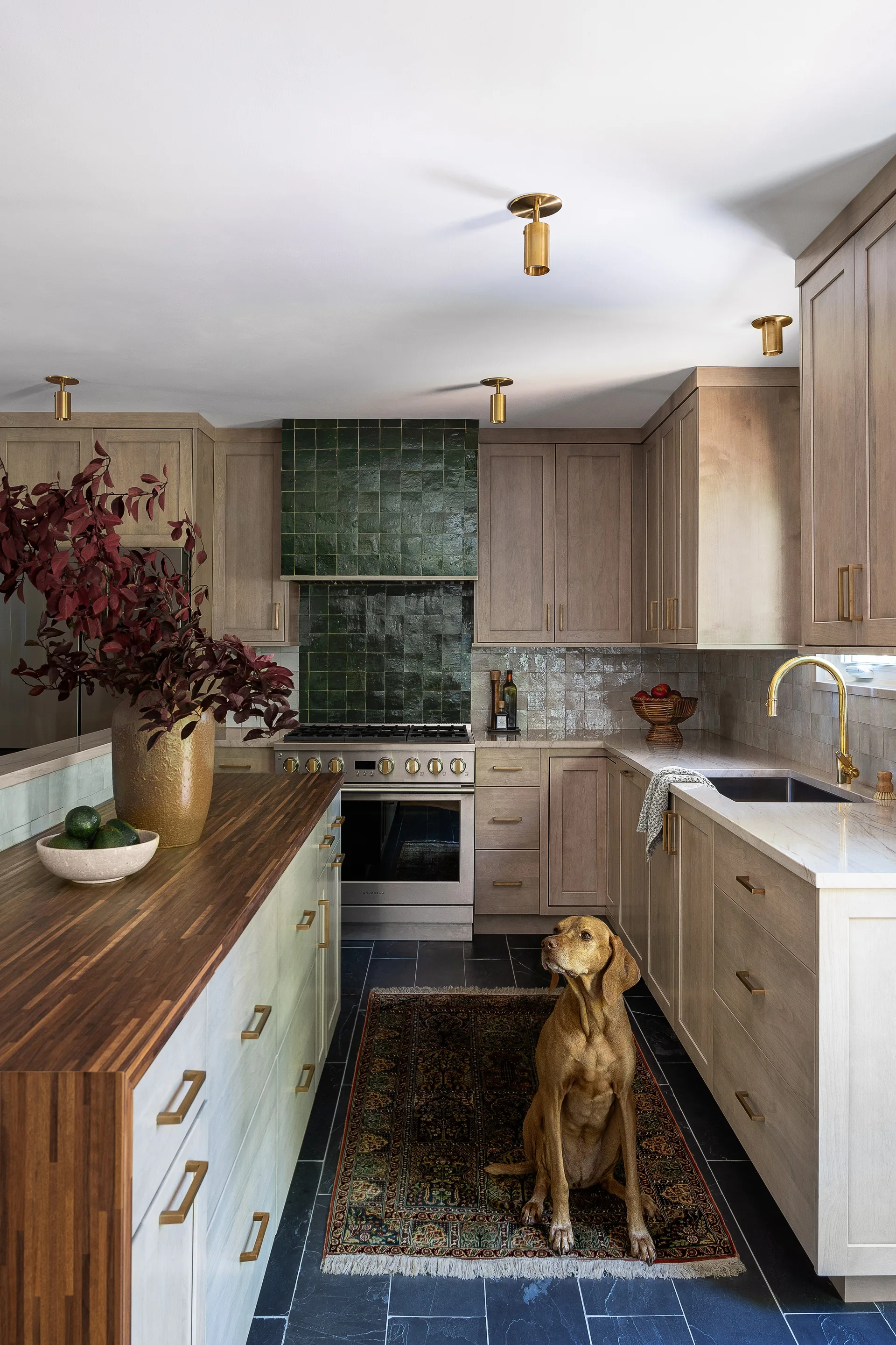 A kitchen features light wood cabinets, a dark green tiled range hood, a butcher block island, and dark slate floor tiles.