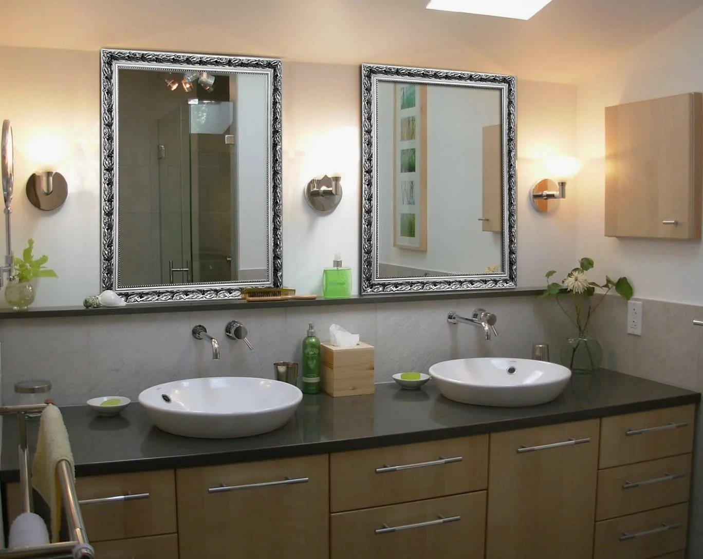 A modern double-sink bathroom vanity with light wood cabinets, dark grey countertop, and two ornate silver-framed mirrors.