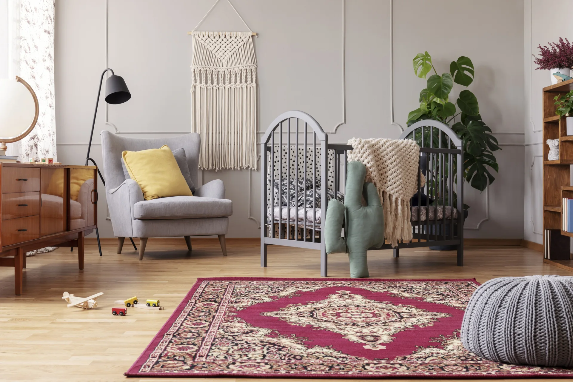 A light grey nursery featuring an arched grey crib, a wooden sideboard, a wingback armchair, and a burgundy patterned rug on hardwood floors.