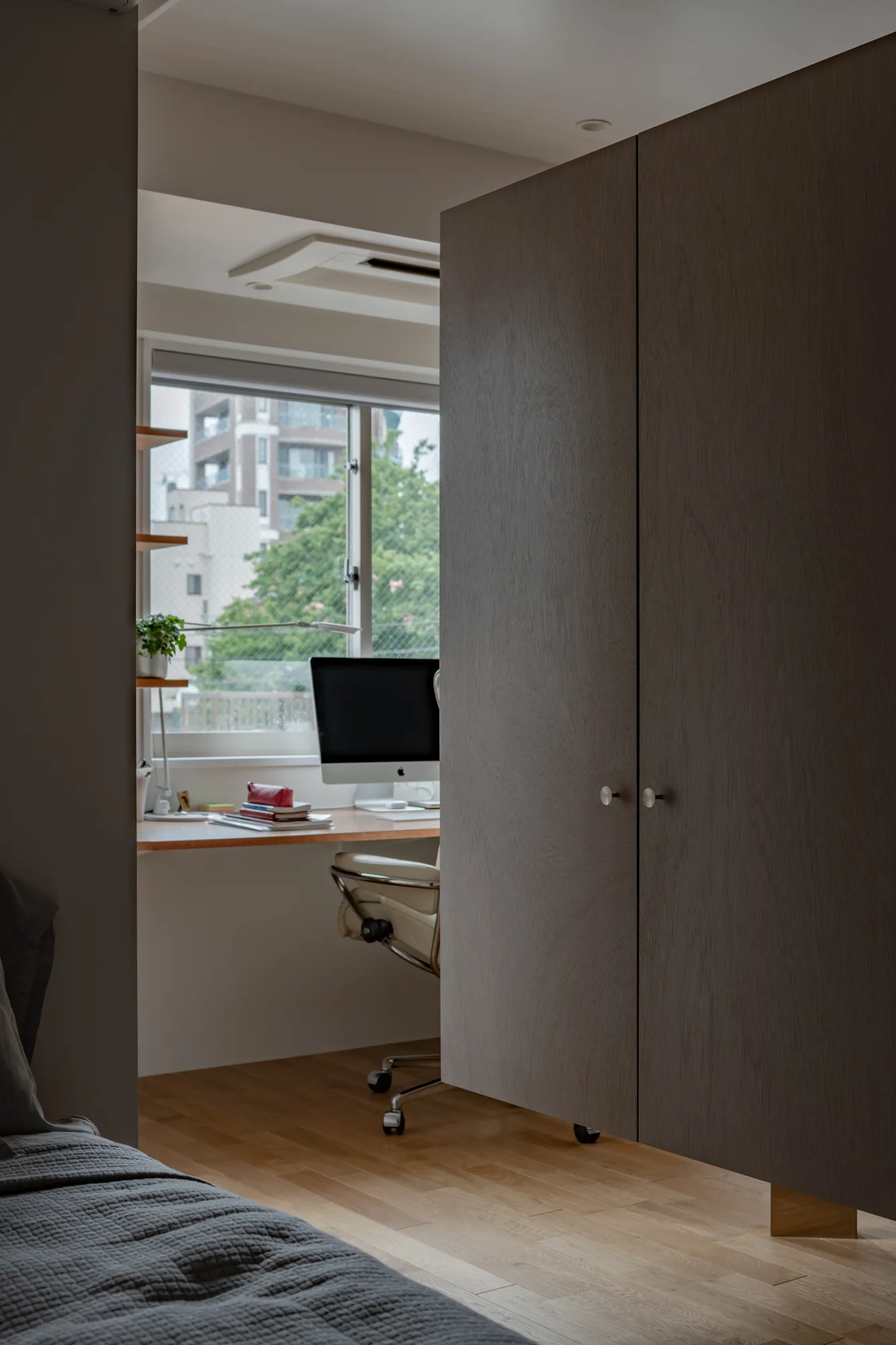 A home office setup with a wood desk, desktop computer, and white office chair next to a tall grey-brown wardrobe cabinet.