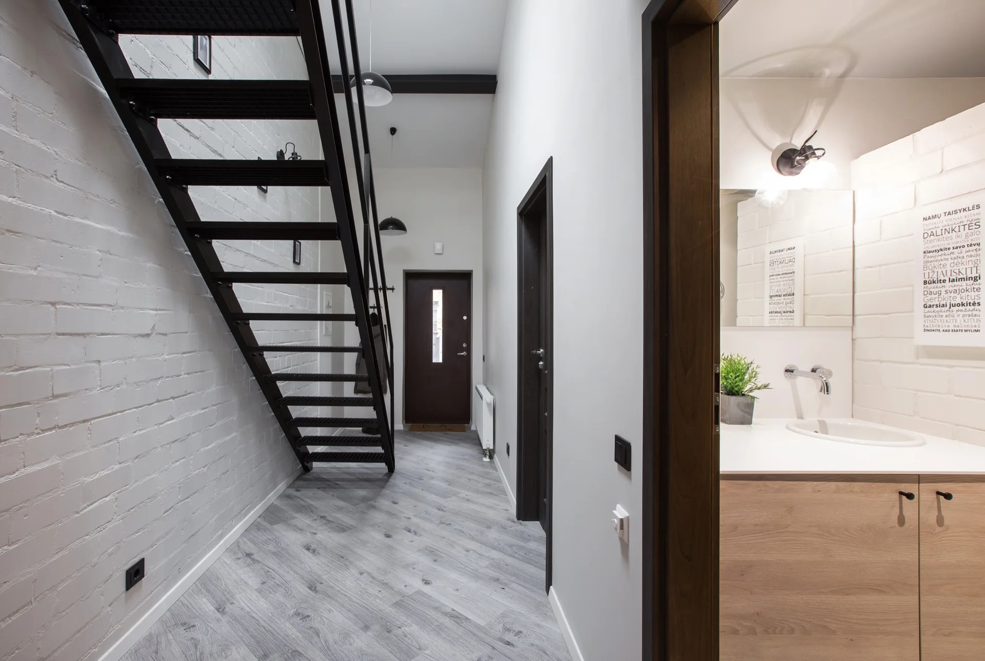 A modern entryway features a black metal open-tread staircase against a white painted brick wall, with a view into an adjacent bathroom.